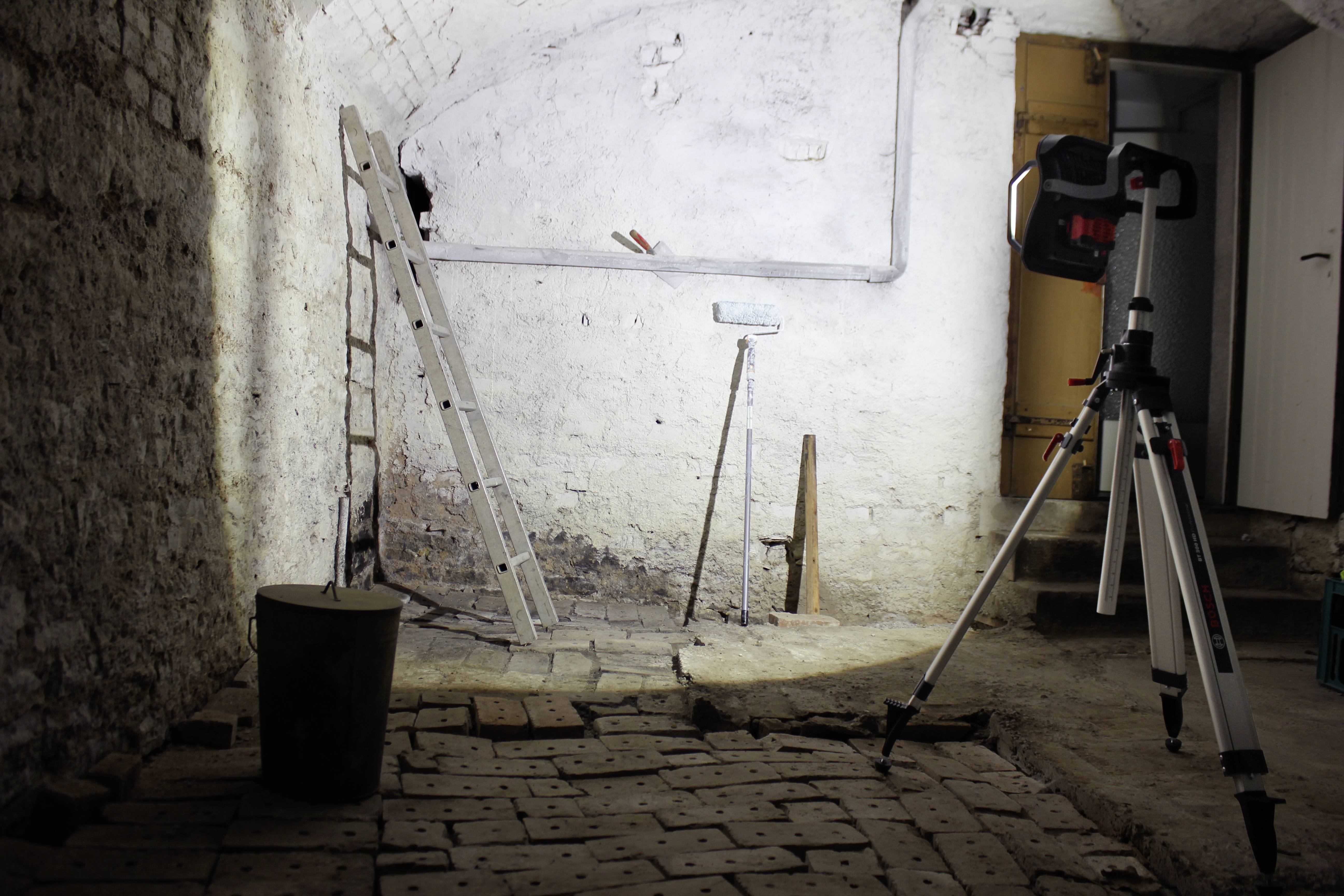 Egy pinceszoba téglapadlóval, egy létrával a falon, egy vödörrel és egy állvánnyal. A háttér gyengén megvilágított.
