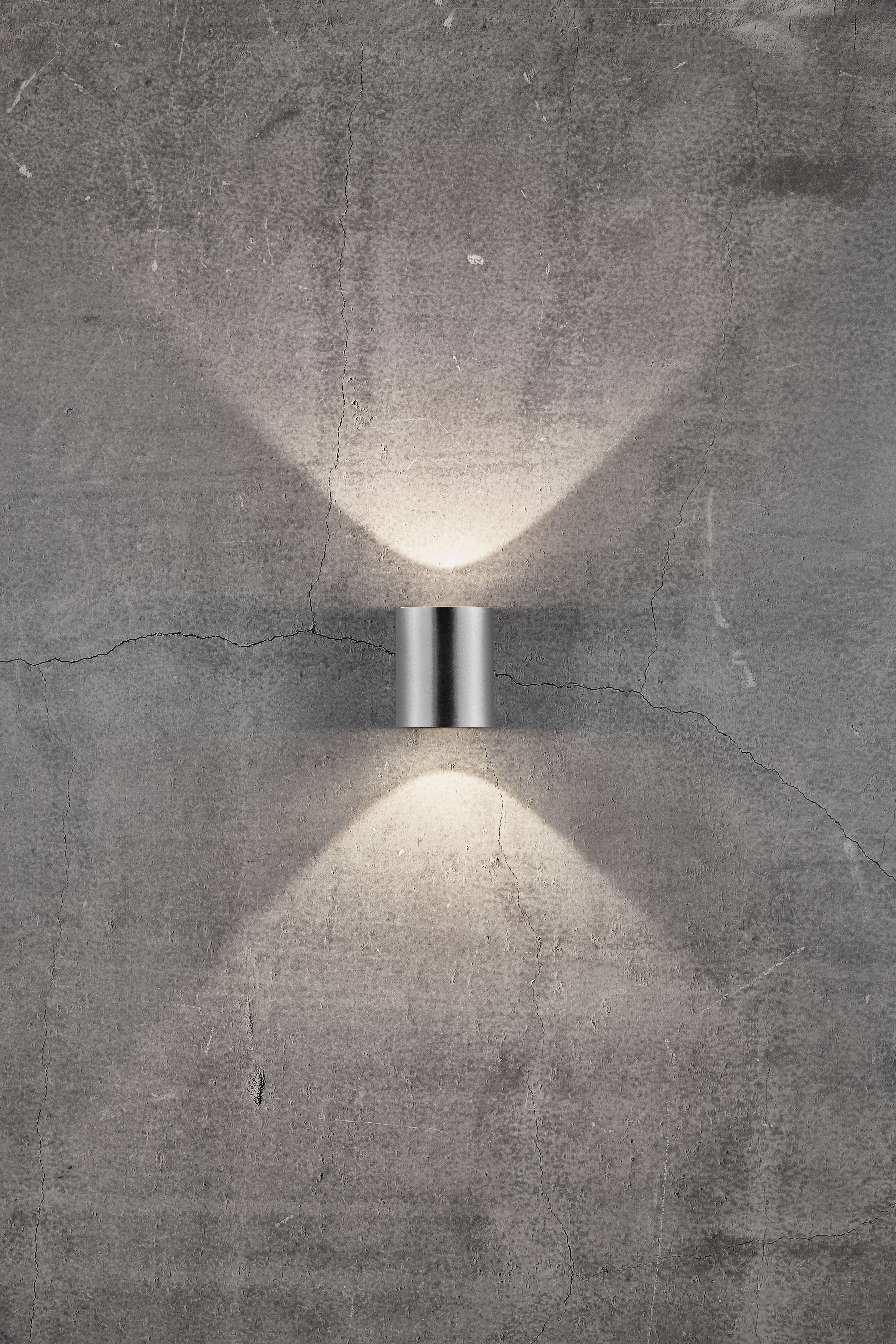 Egy modern fali lámpa van felszerelve egy szürke betonfalra. Felfelé és lefelé sugároz fényt, és lágy, egyenletes mintázatot hoz létre.