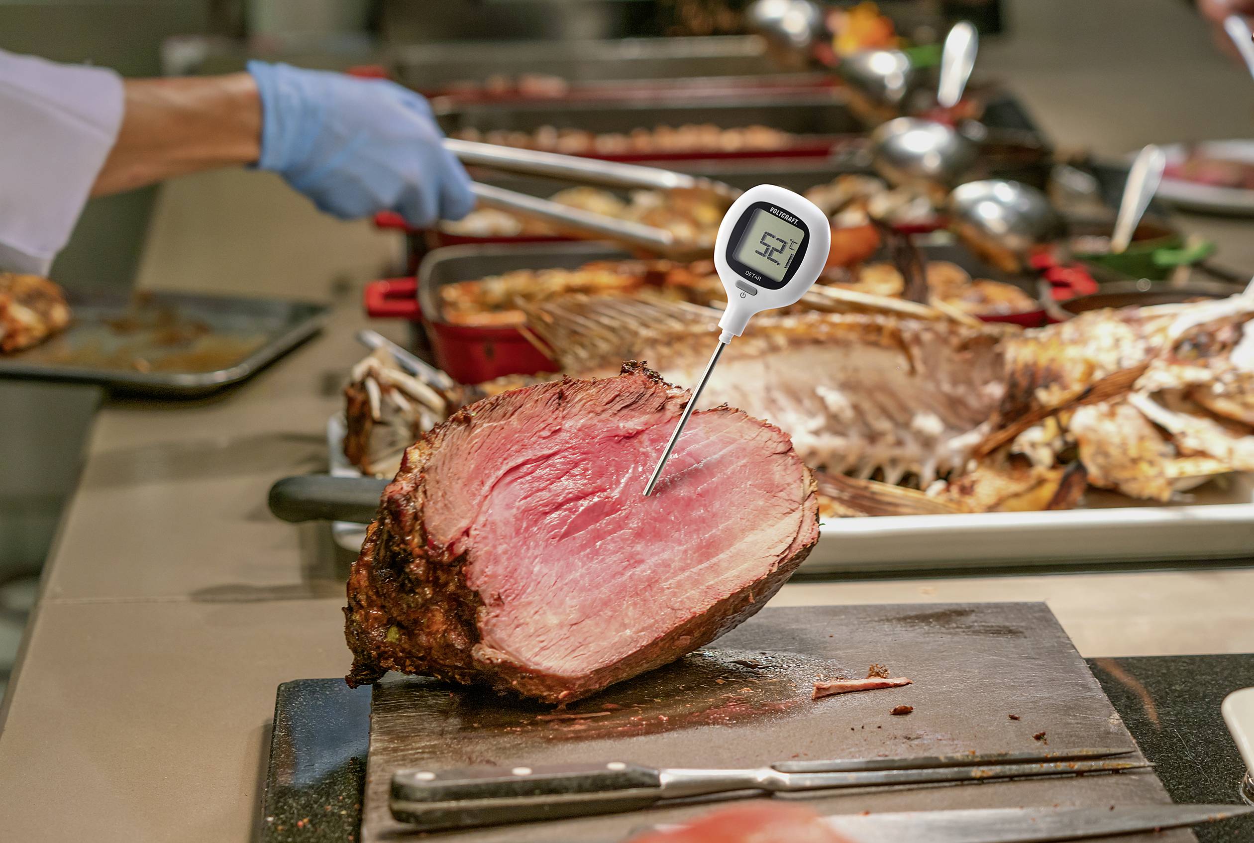 Egy nagy darab sült húst egy hőmérővel ellenőriznek, amely 52 fokot mutat. A háttérben további húsételek láthatók.