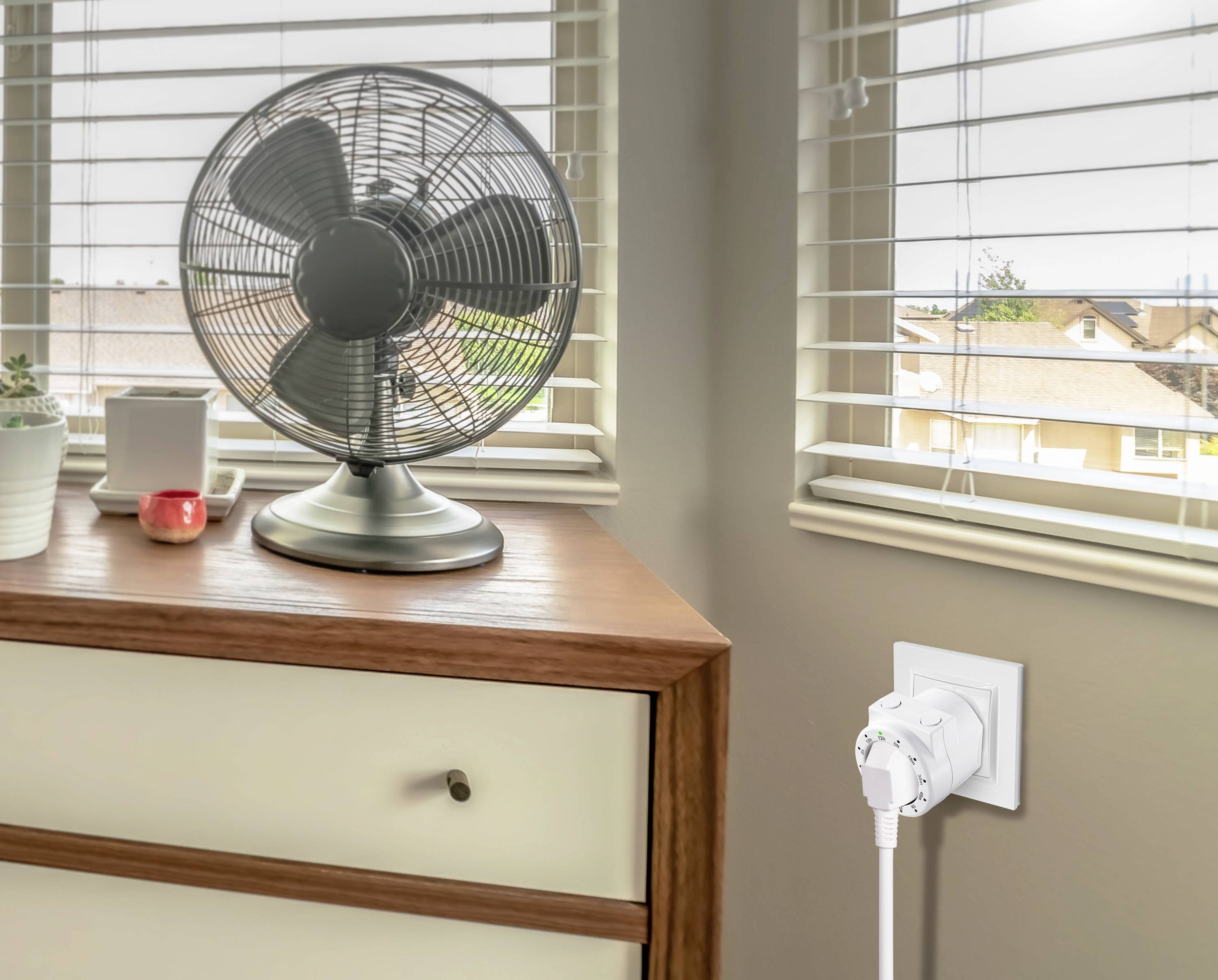 Egy asztali ventilátor áll egy komódon. A háttérben ablakok láthatók redőnyökkel, amelyek beengedik a nappali fényt.