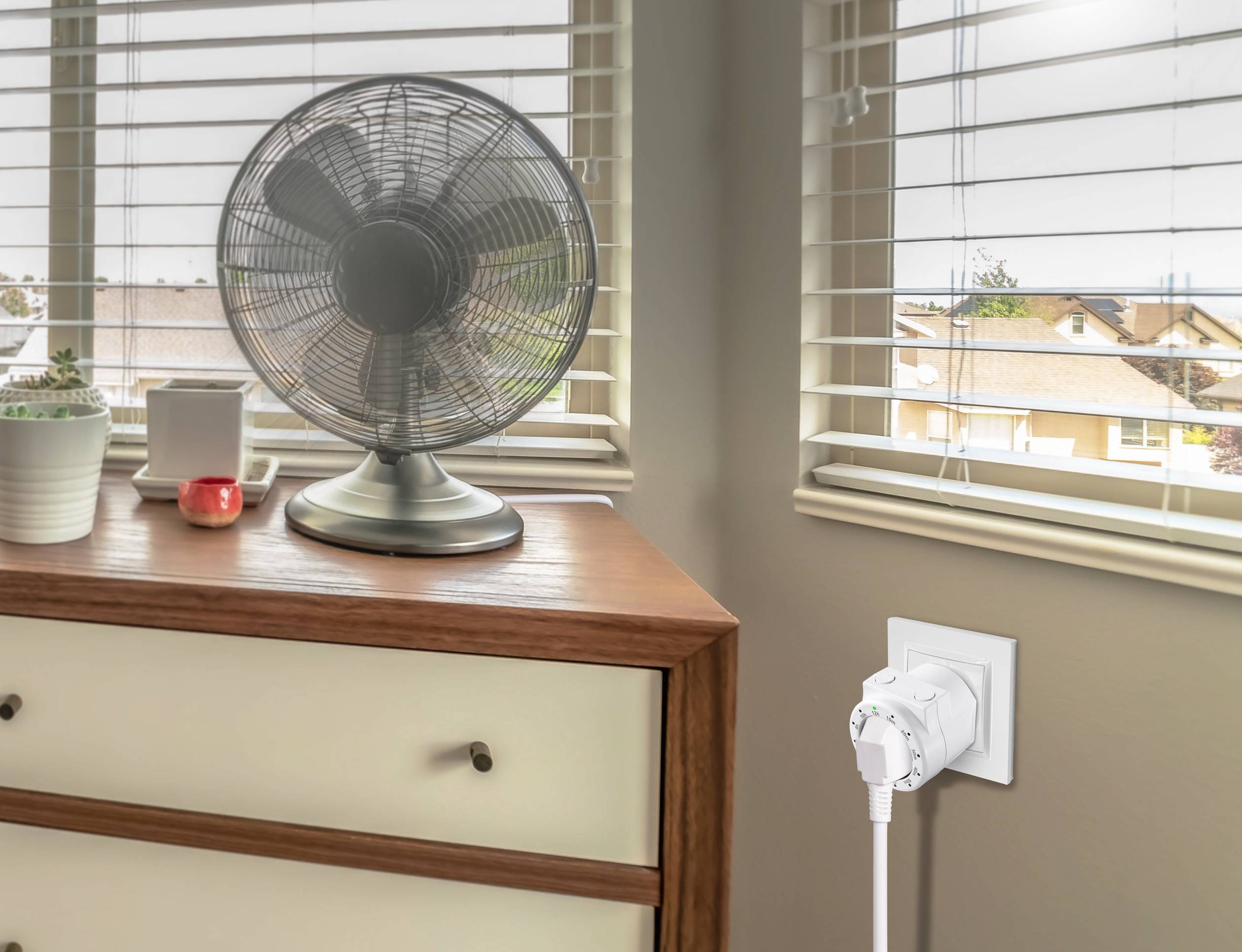 Egy ventilátor áll egy fiókos szekrényen egy világos helyiségben. Egy okosotthon eszköz van csatlakoztatva a fali konnektorba.