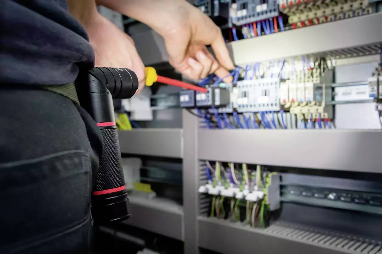 'Egy személy egy csavarhúzóval elektromos kapcsolótáblát javít, miközben egy elemlámpa lóg az övén.'