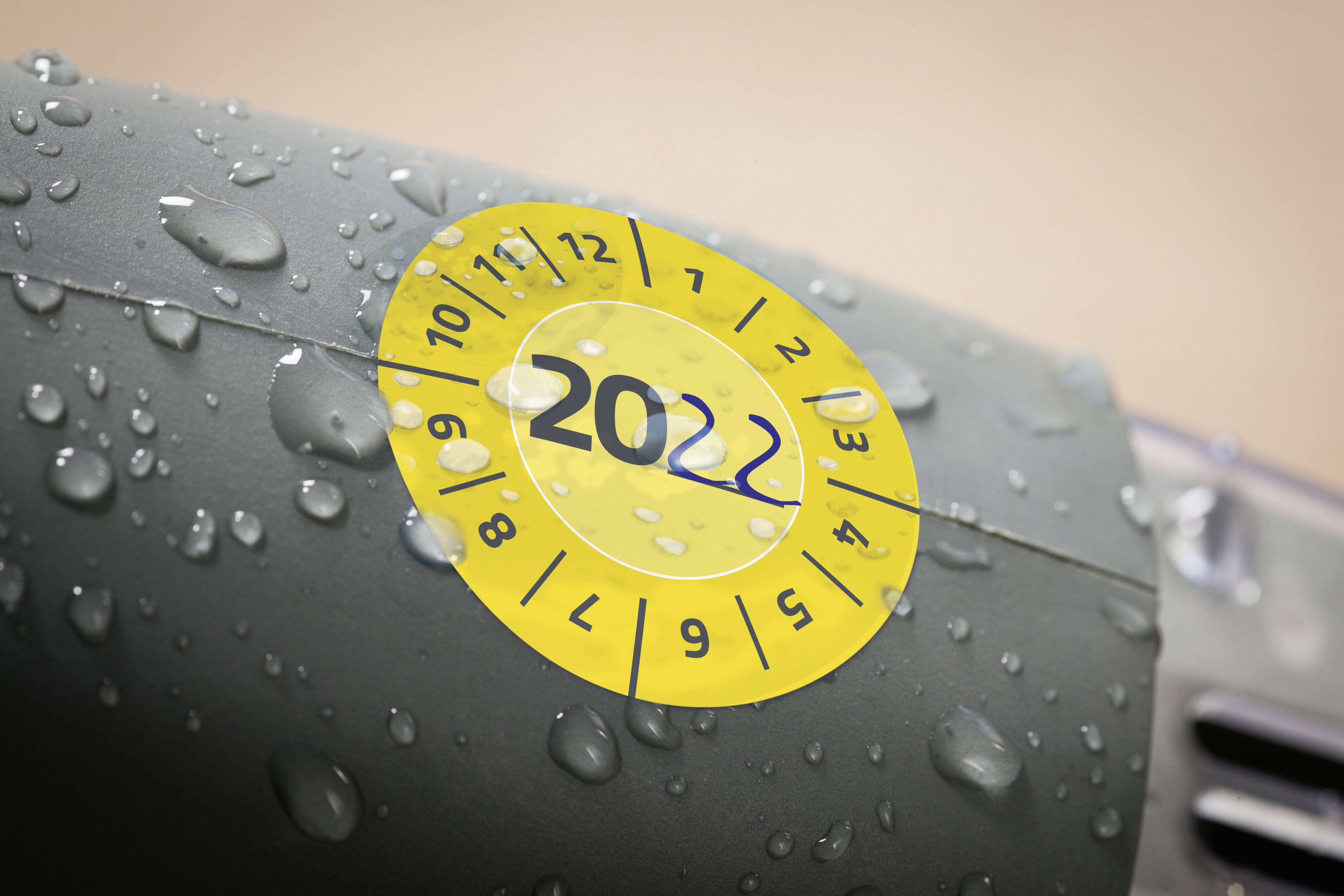Sárga TÜV-matrica a '22' számmal egy autón, amely a 2022-es évre esedékes műszaki vizsgát jelzi. Esőcseppek vannak rajta.