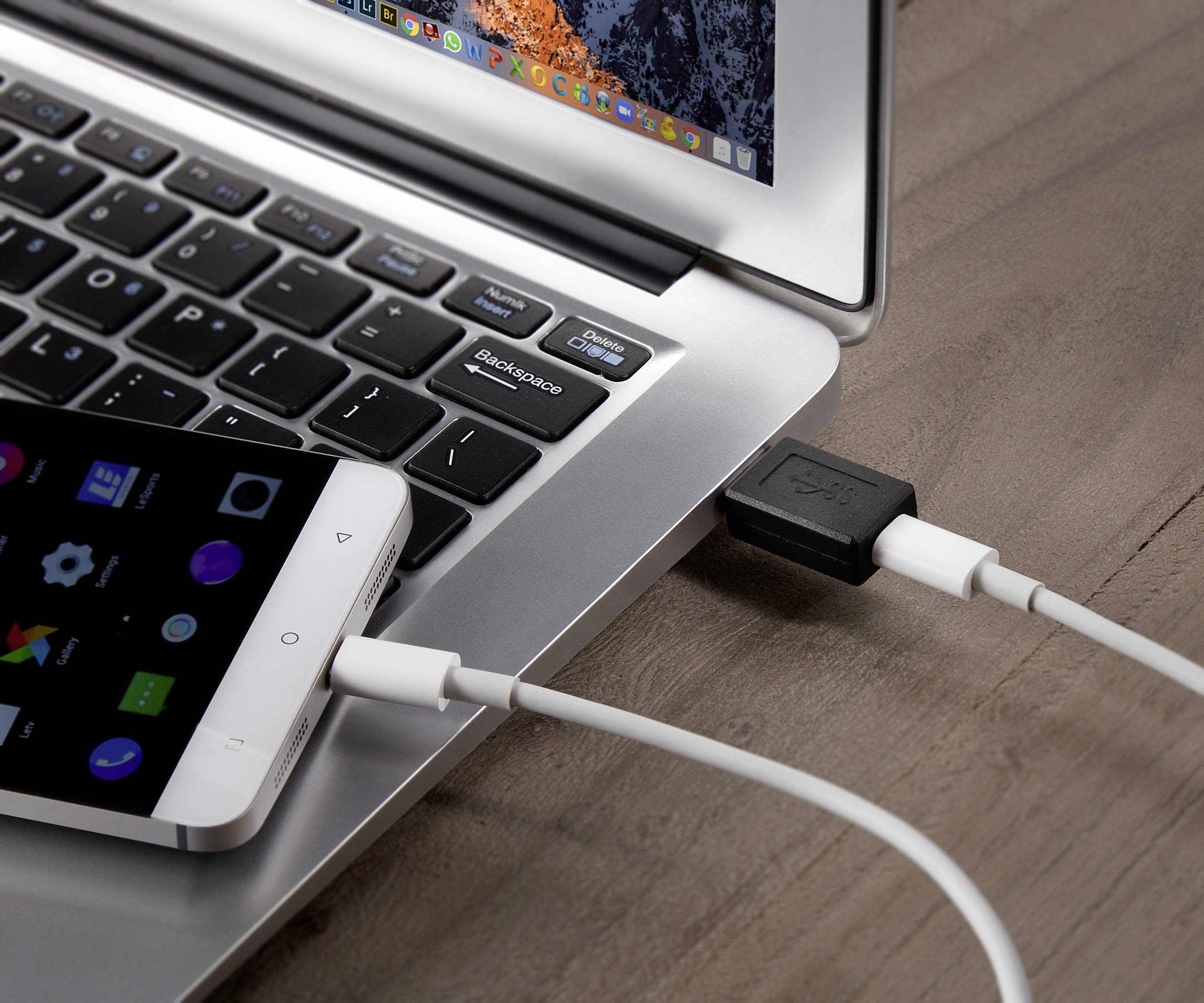 Egy mobiltelefon USB-kábellel csatlakoztatva egy laptophoz, fából készült asztalon látható. A laptop képernyőjén színes ikonok jelennek meg.