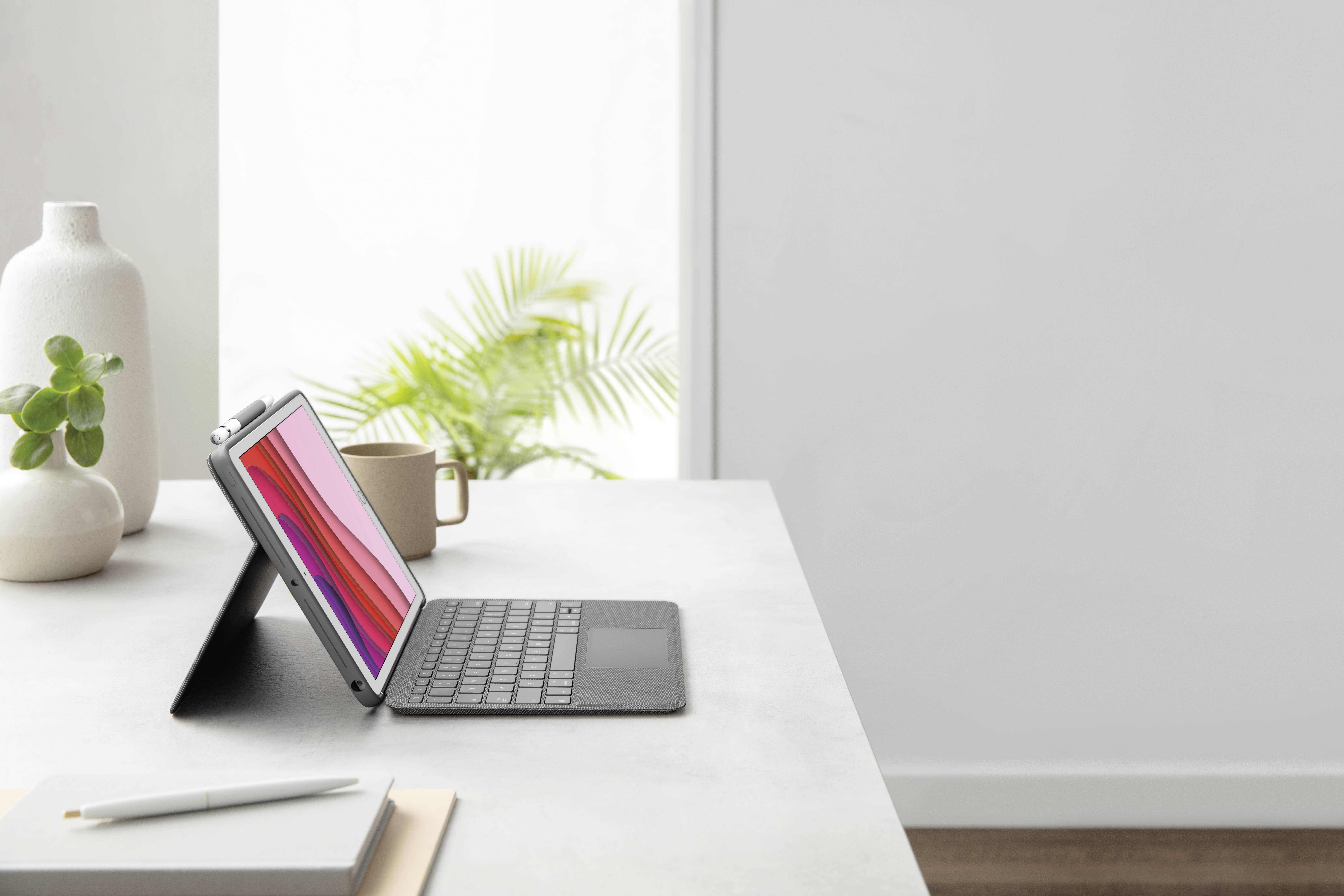 Egy tablet billentyűzetes tokkal egy íróasztalon, kivilágított képernyővel, egy kávéscsésze és egy növény mellett. Világos, minimalista tér.