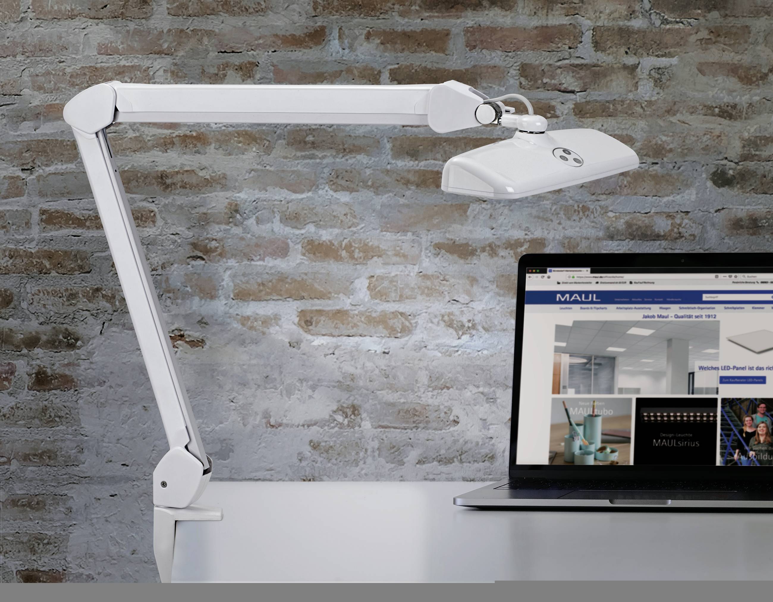 Egy fehér, csuklós karú íróasztali lámpa áll egy asztalon egy laptop mellett egy téglafal előtt. A laptop egy weboldalt jelenít meg.