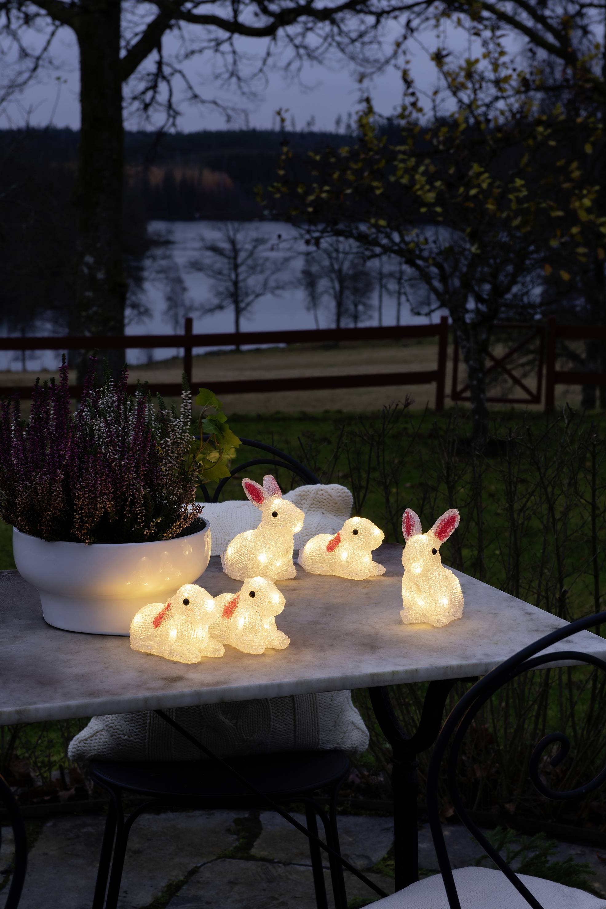 Alkonyatkor világító nyúlfigurák egy asztalon, növények között. A háttérben egy tó és egy fa. Hangulatvilágítás a kertben.