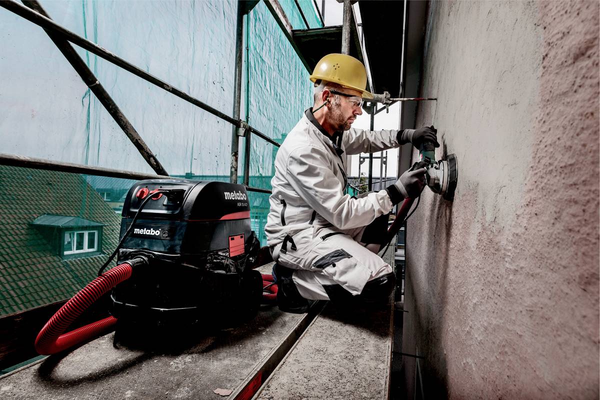 Egy munkás védősisakban és kesztyűben csiszol egy falat egy állványon. Mellette egy porszívó áll.