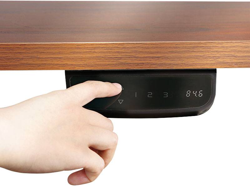 Egy kéz egy digitális vezérlőgombot nyom egy fa asztal alatt, amelynek magasságállítási lehetősége és kijelzője '84.6' értéket mutat.