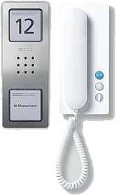 Lakás falán lévő kaputelefon kijelzővel, amelyen a 'M. Mustermann' név szerepel, kommunikációs gombokkal és fehér telefonfejhallgatóval.