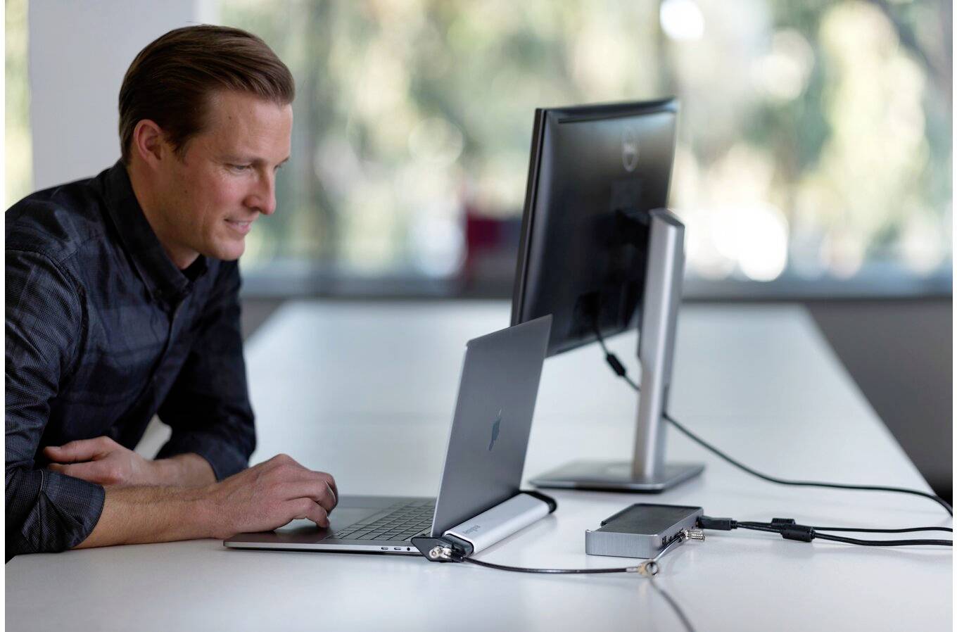 Egy férfi egy laptopon dolgozik, amely egy külső monitorhoz csatlakoztatva van egy világos irodában. Teljes figyelmét a képernyőre összpontosítja.