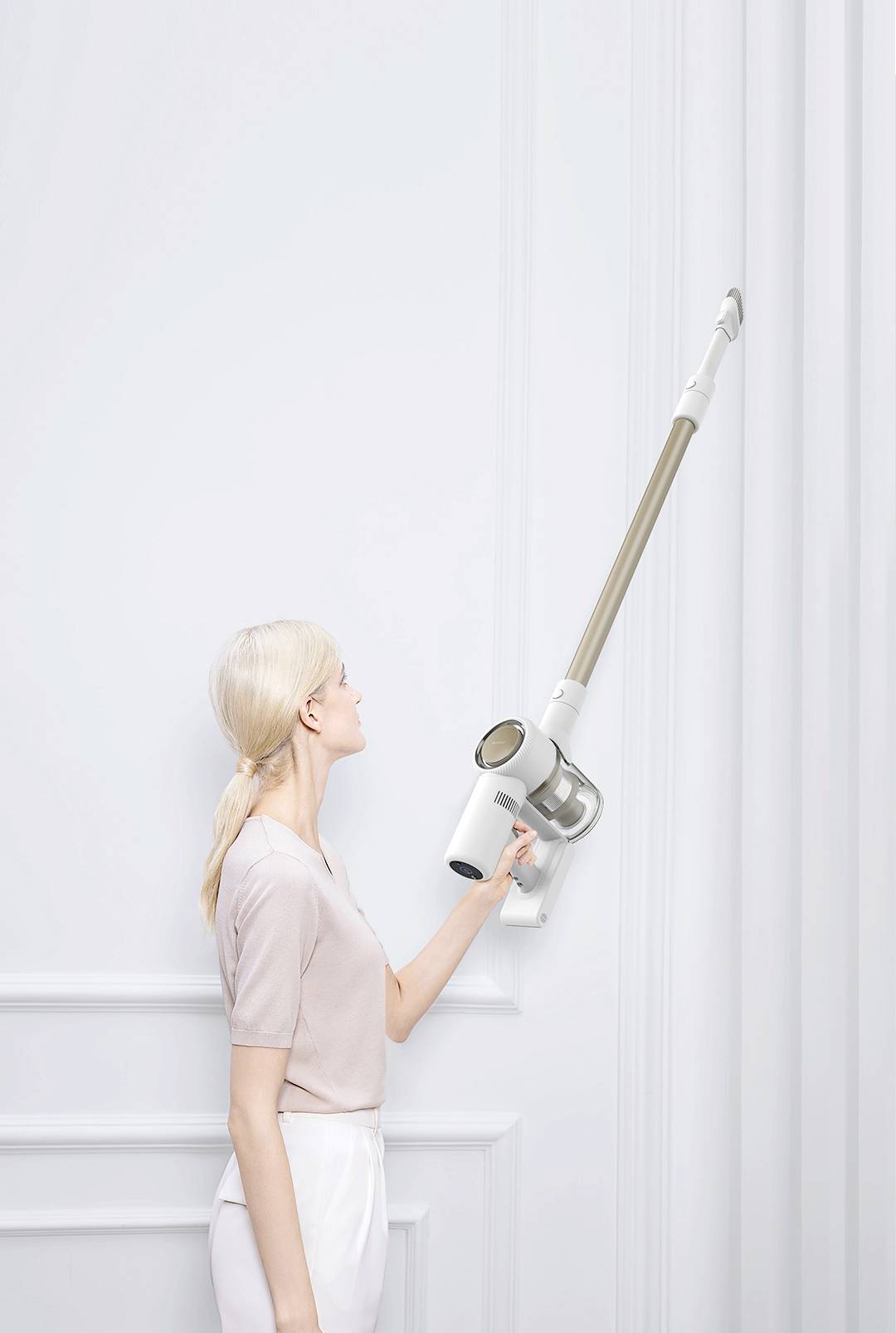 Egy nő egy vezeték nélküli porszívóval takarít egy fehér falat egy minimalista helyiségben.