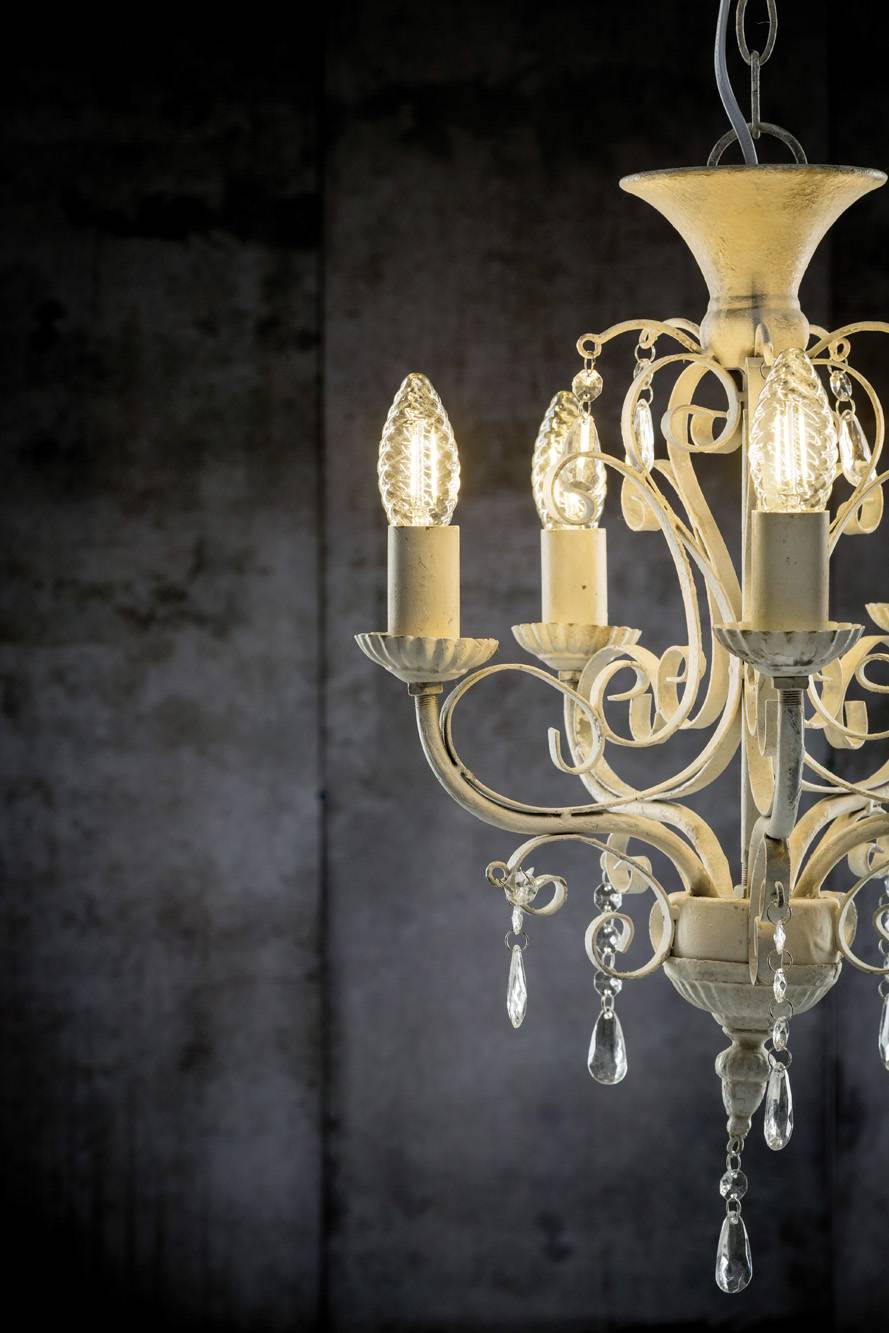 Egy fehér csillár dekoratív részletekkel és öt világító gyertyalámpával sötétszürke háttér előtt.