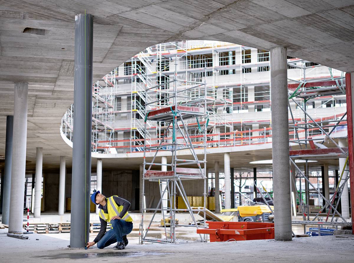 Egy sárga biztonsági mellényes építőmunkásnő ellenőriz egy betonoszlopot egy állványokkal körülvett és befejezetlen épület hátterében.