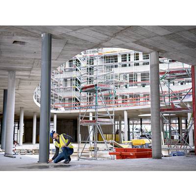 Egy sárga biztonsági mellényes építőmunkásnő ellenőriz egy betonoszlopot egy állványokkal körülvett és befejezetlen épület hátterében.
