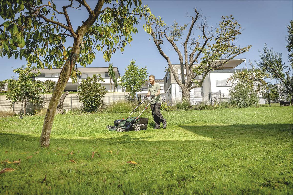 Egy férfi füvet nyír egy napsütéses napon egy kertben, amelyet fák és modern lakóépületek vesznek körül a háttérben.