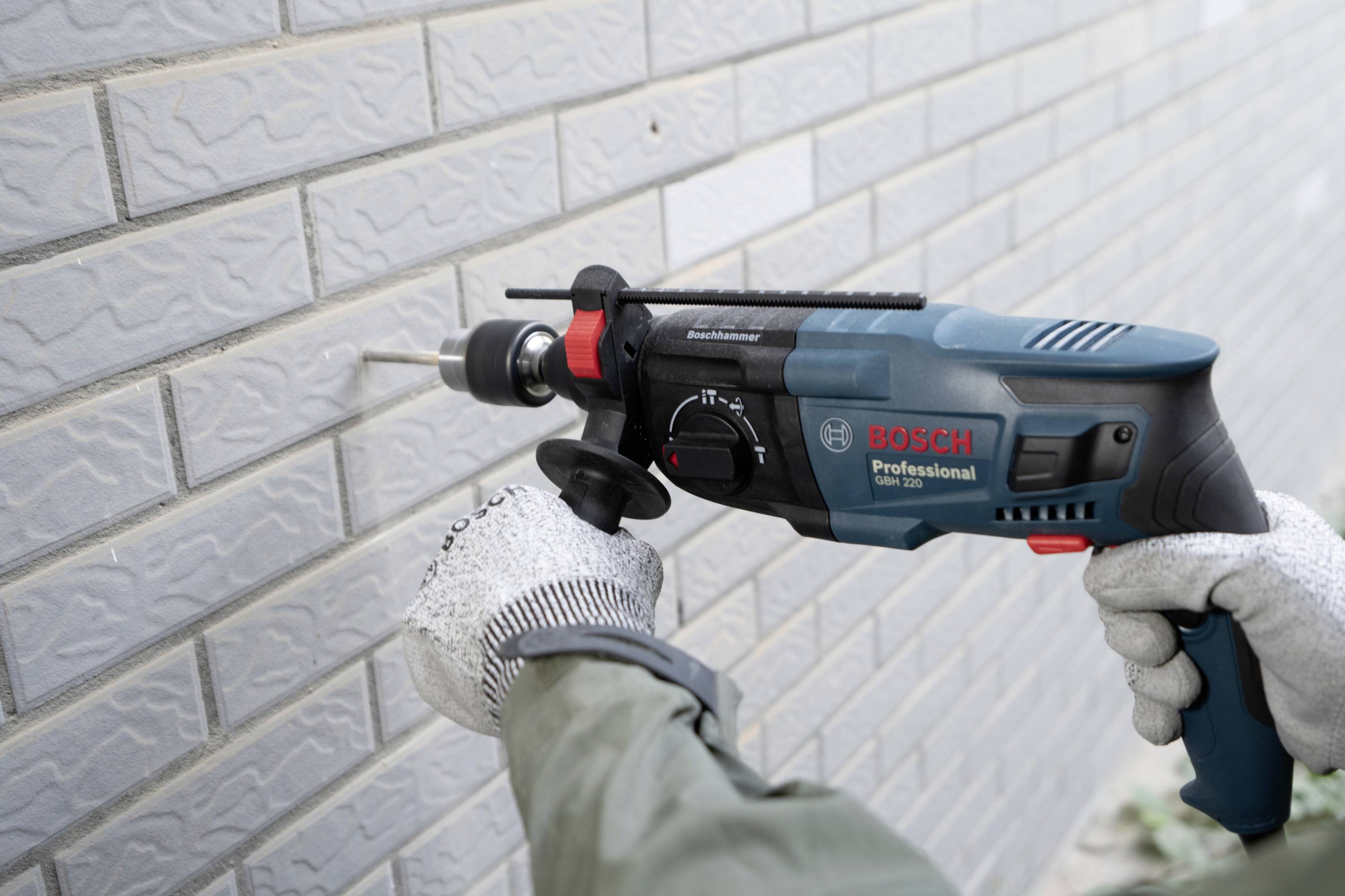Egy személy egy Bosch Professional fúrókalapáccsal lyukat fúr egy téglafalba, miközben kesztyűt és kabátot visel.