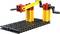 Egy LEGO daru modell két oldalsó hajtókarral. Egy sárga rúd köti össze a két piros tornyot egy fekete alaplapon.
