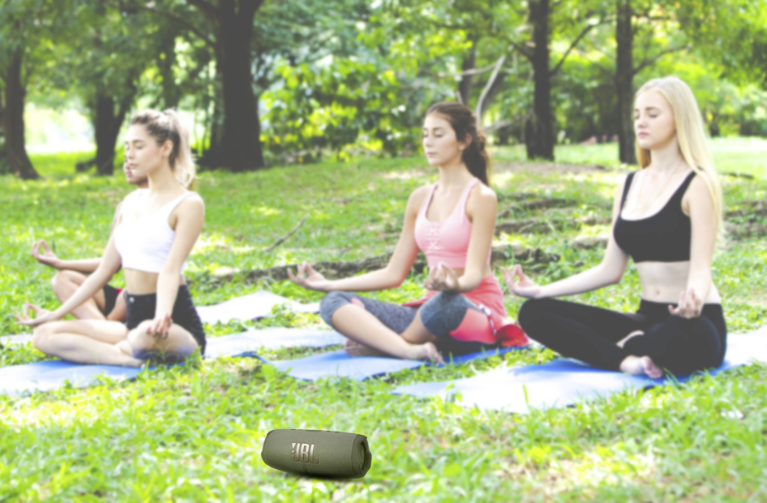 Három személy meditál jógaszőnyegeken egy zöld parkban, szabadtéren. Törökülésben ülnek, csukott szemmel.