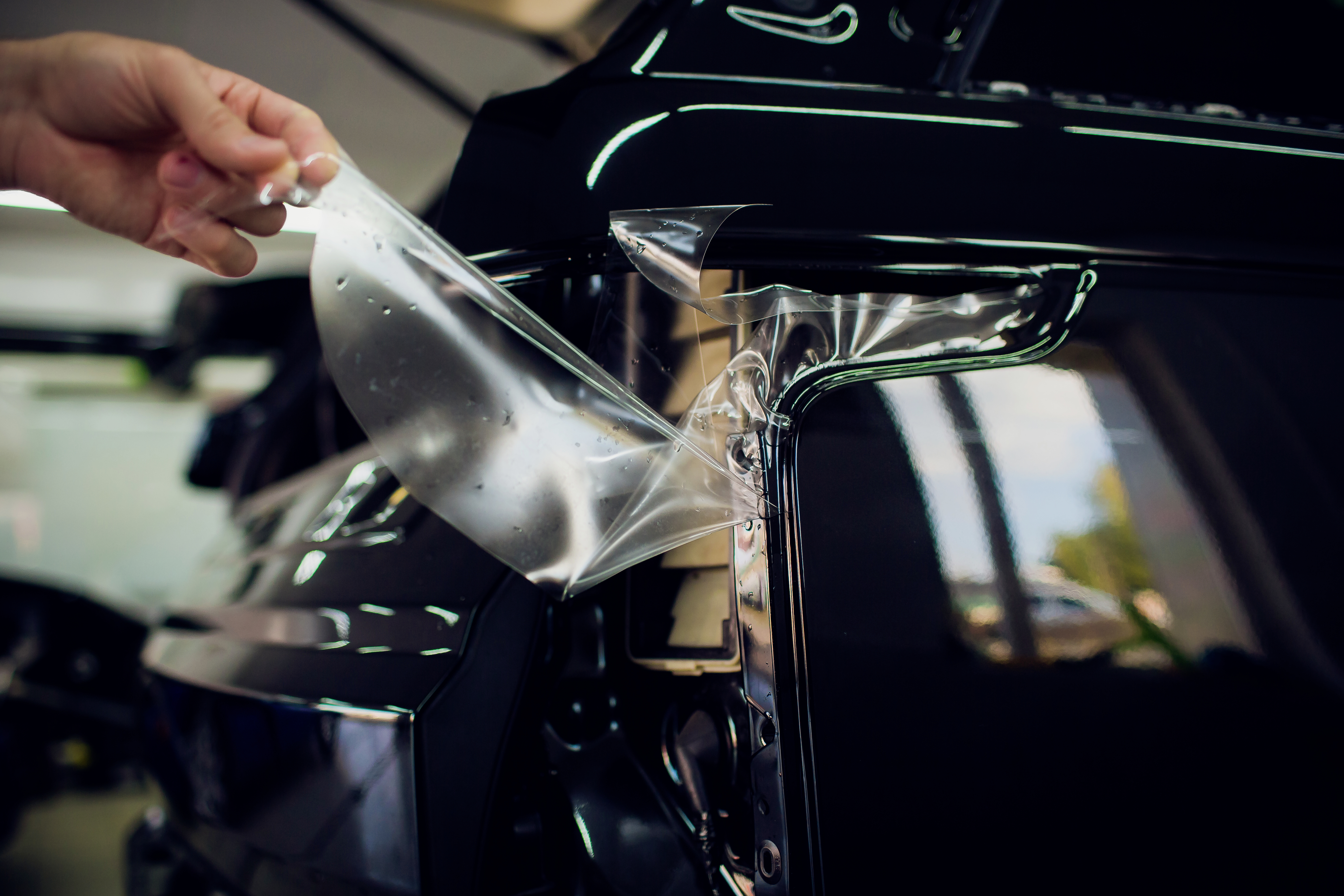 Egy kéz lehúz egy védőfóliát egy fényes fekete autóalkatrészről, hogy feltárja vagy megvédje a felületet.