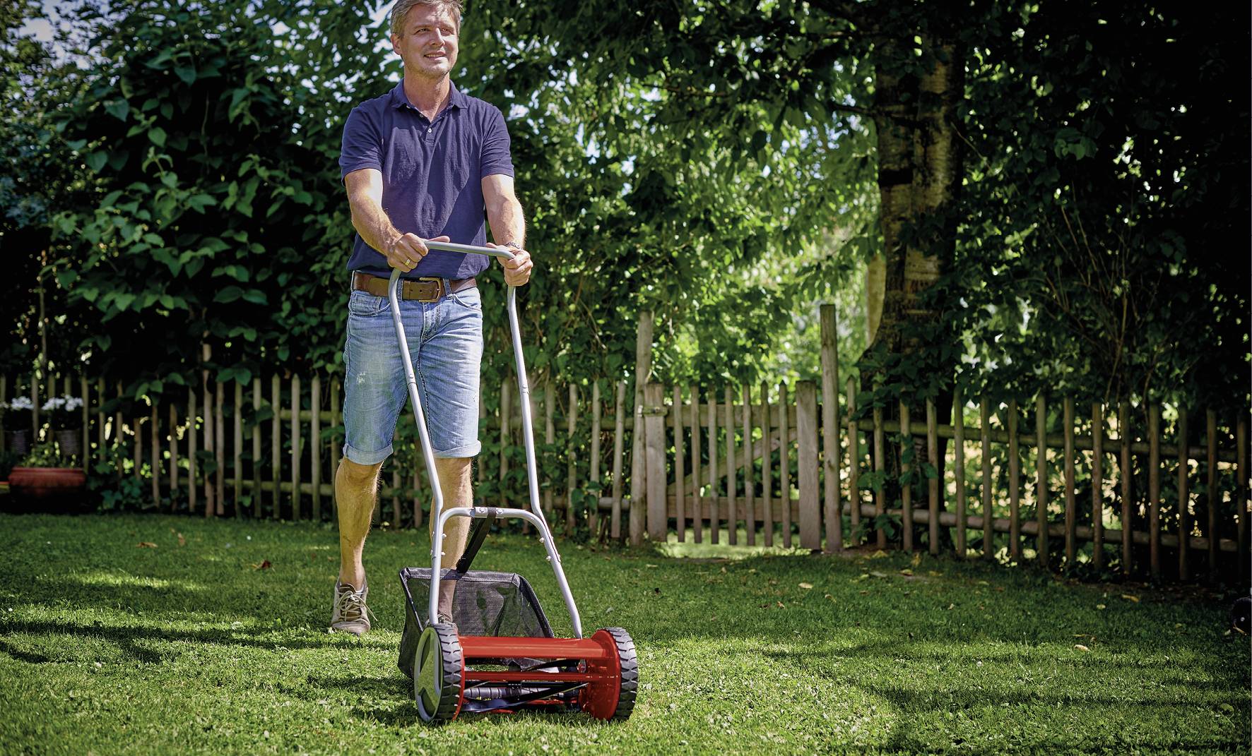 Egy kék pólós és farmershortot viselő személy egy kézi tologatós fűnyíróval nyírja a füvet egy zöld kertben.