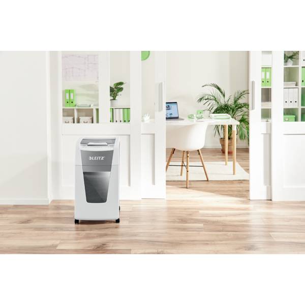 'Iroda világos fa padlóval és fehér polcokkal, előtérben egy iratmegsemmisítő, háttérben egy íróasztal laptoppal.'