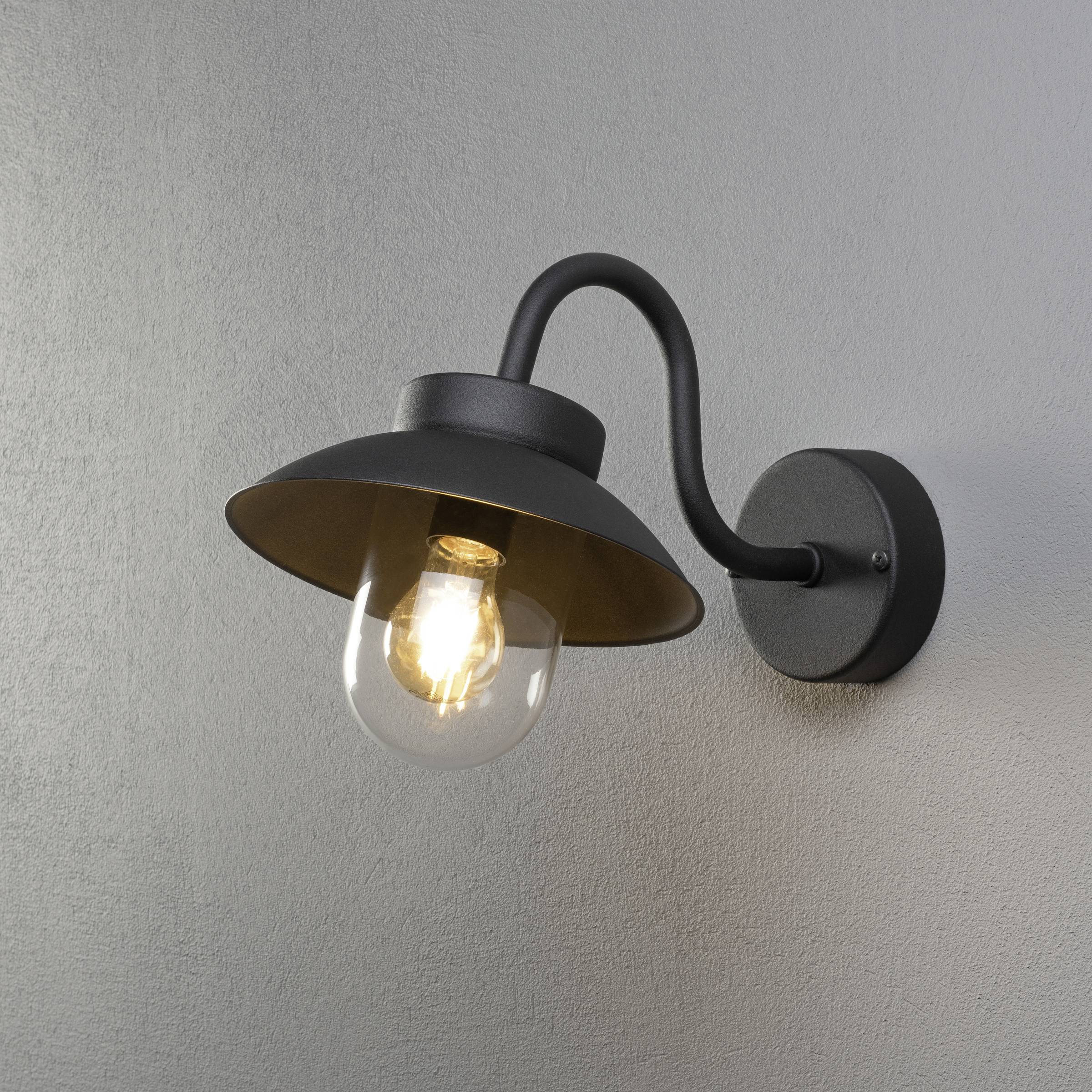 Fekete fali lámpa hajlított karral, szürke falra szerelve, bekapcsolt izzóval kerek üvegburában.