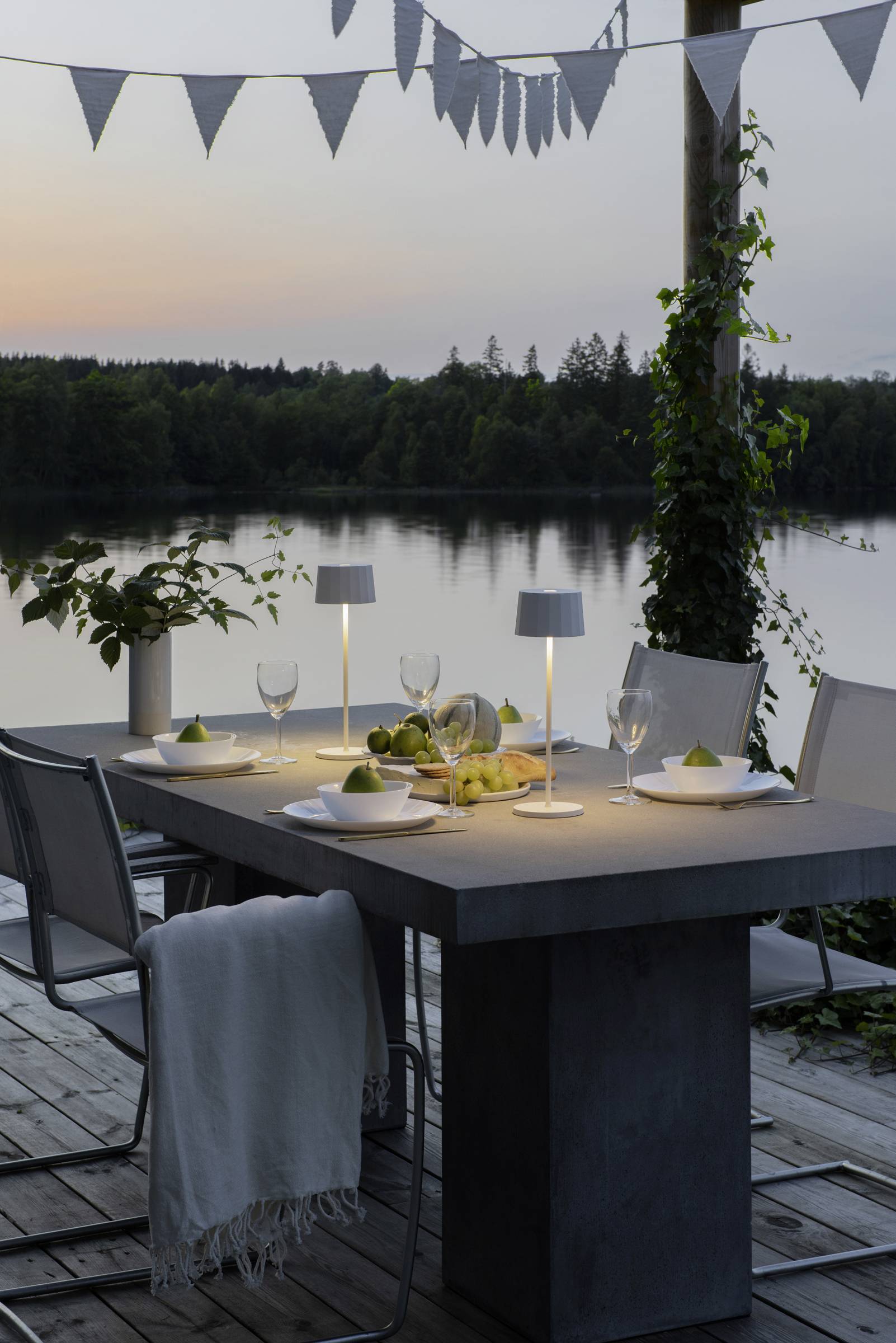 Egy asztal egy teraszon, naplemente idején, megterítve vacsorához, mécsesekkel és tányér-elrendezésekkel, kilátással egy tóra.