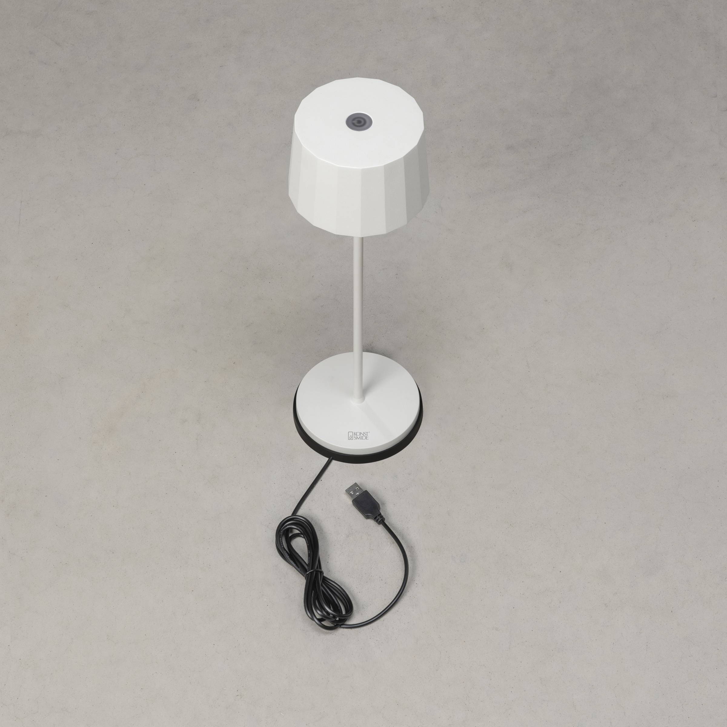 Egy fehér asztali lámpa USB-kábellel szürke háttér előtt. A lámpának kerek talpa és bordázott ernyője van.