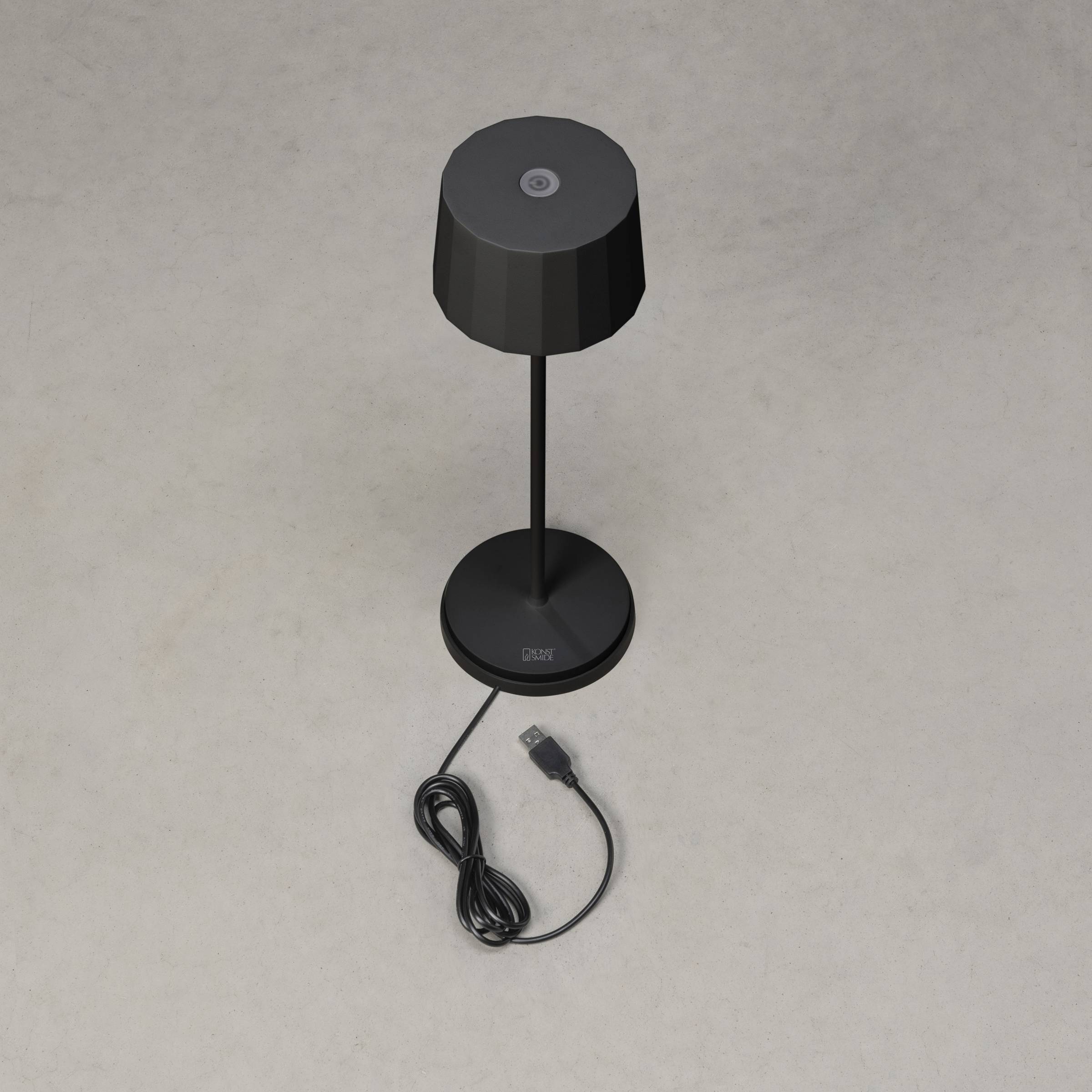 Fekete asztali lámpa USB-csatlakozóval, szürke háttér előtt. A kábel lazán van feltekeredve a lámpa talapzata körül.