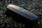 Egy fekete technikai eszköz fekszik a kavicson. Kerek gombbal rendelkezik és a 'CTEK' logóval van ellátva. Valószínűleg egy töltőkészülékről van szó.