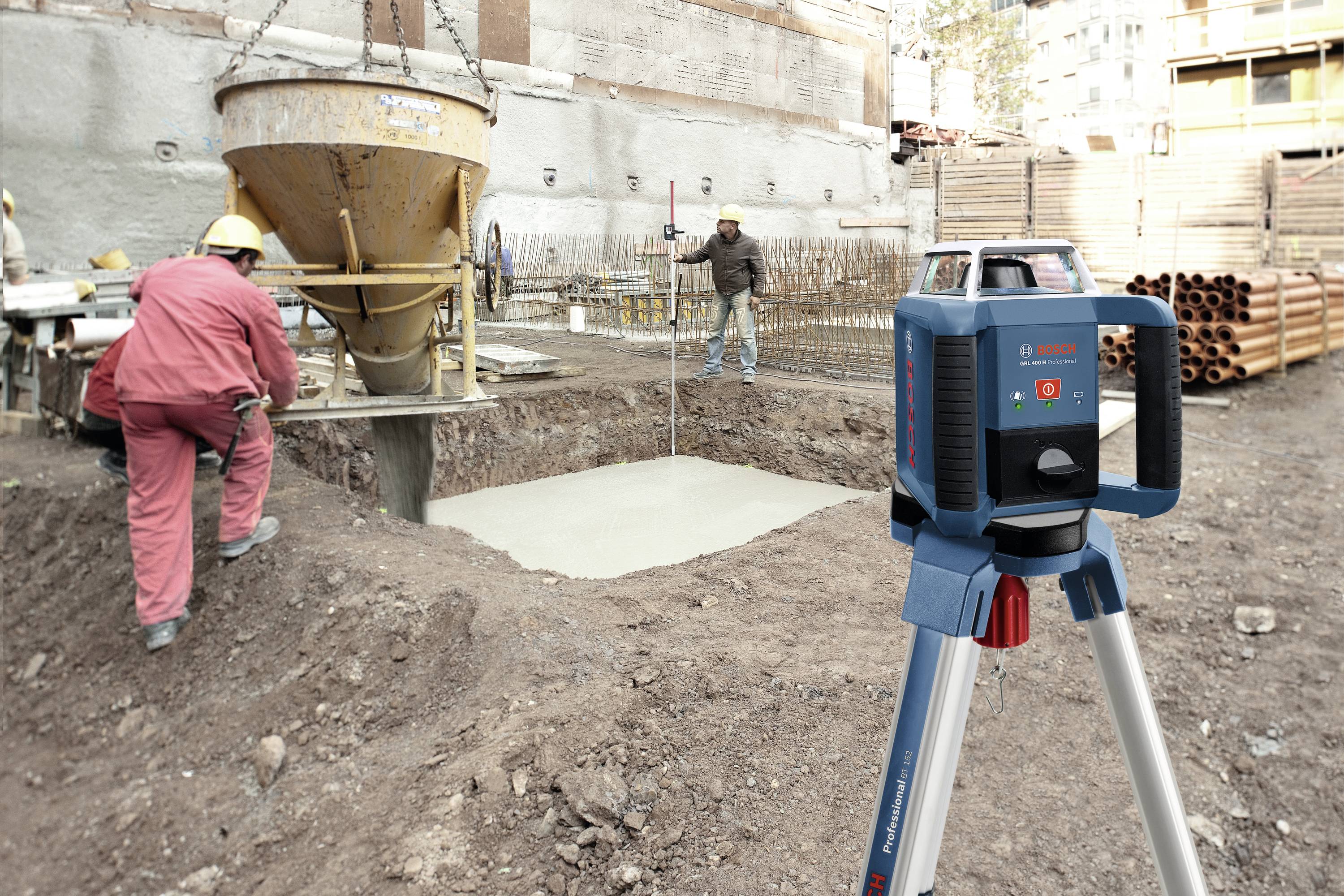Munkások betonnal töltenek fel egy alapzatot, miközben az előtérben egy teodolitok áll állványon a felméréshez.