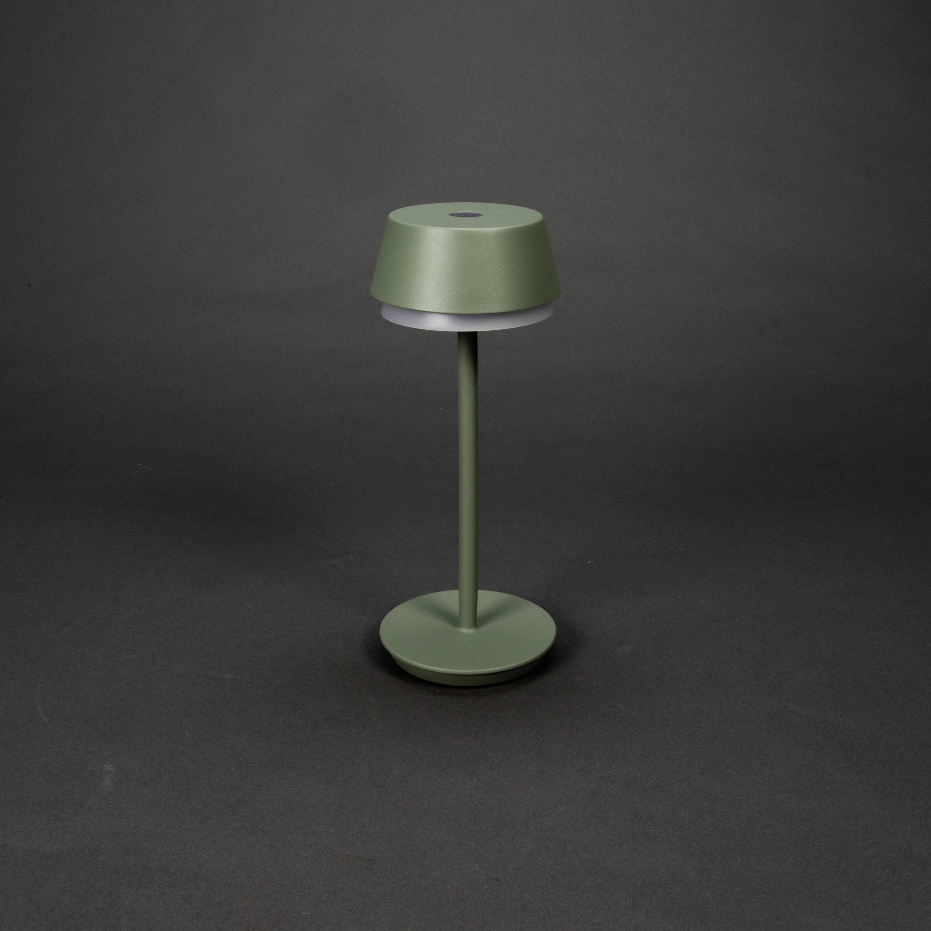 Egy zöld asztali lámpa hengeres ernyővel és egyszerű dizájnnal sötét háttér előtt áll.