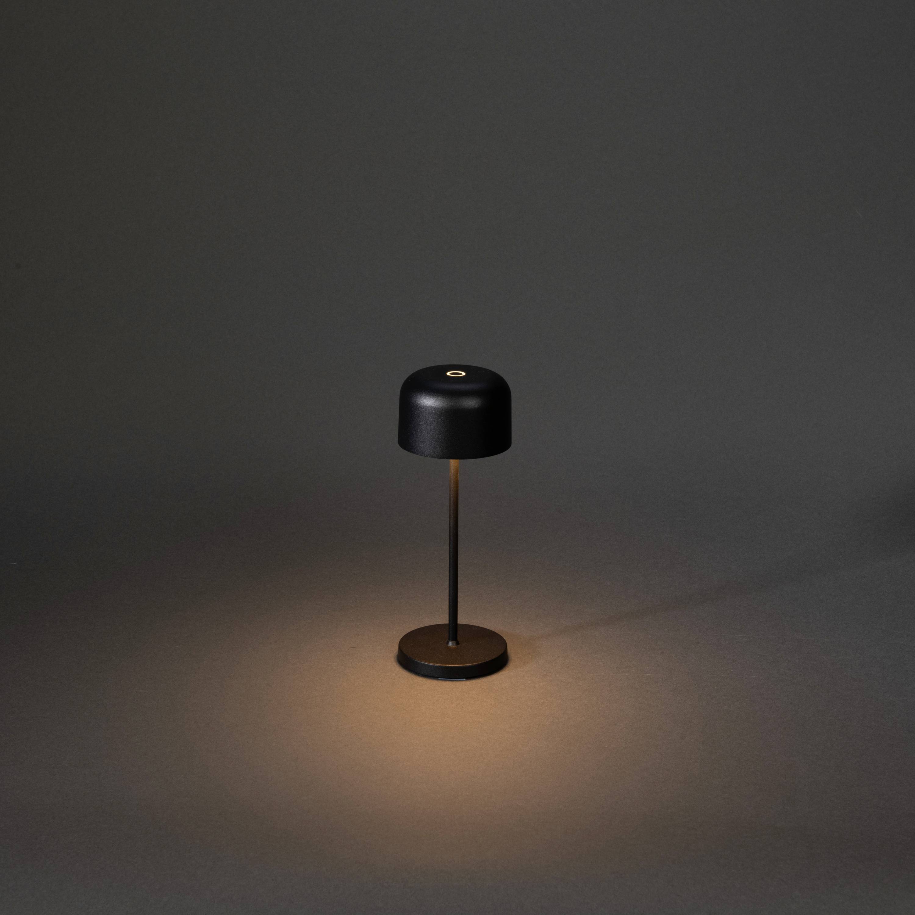 Egy minimalista fekete asztali lámpa az asztalon, amely meleg fényt áraszt egy sötét szobában.