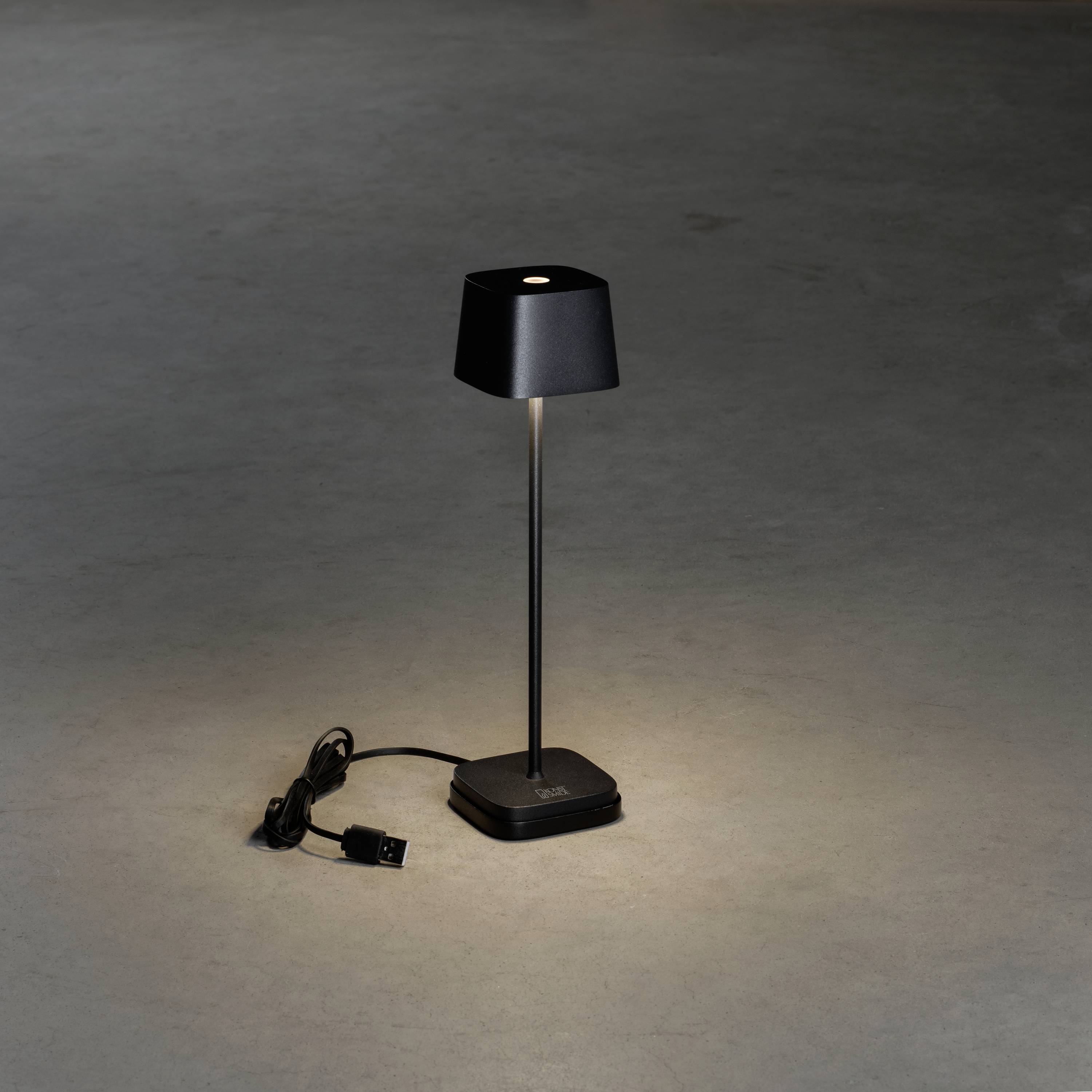 Fekete asztali lámpa keskeny állvánnyal és négyzet alakú ernyővel, szürke padlón áll. A kábel a csatlakozóval mellette fekszik.