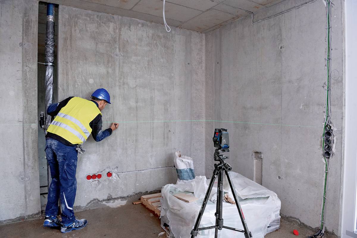 Egy sárga mellényes munkás lézereszközzel méri a betonfalat. A háttérben építkezési terület látható kábelekkel és építőanyagokkal.