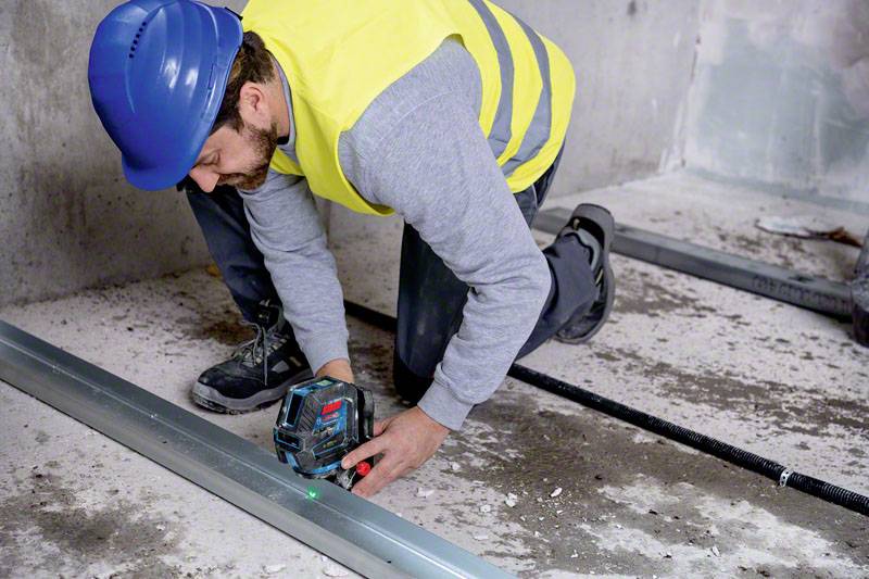Egy munkás kék sisakban és sárga mellényben egy elektromos szerszámmal fémtámaszokat rögzít egy épülő szerkezetben.