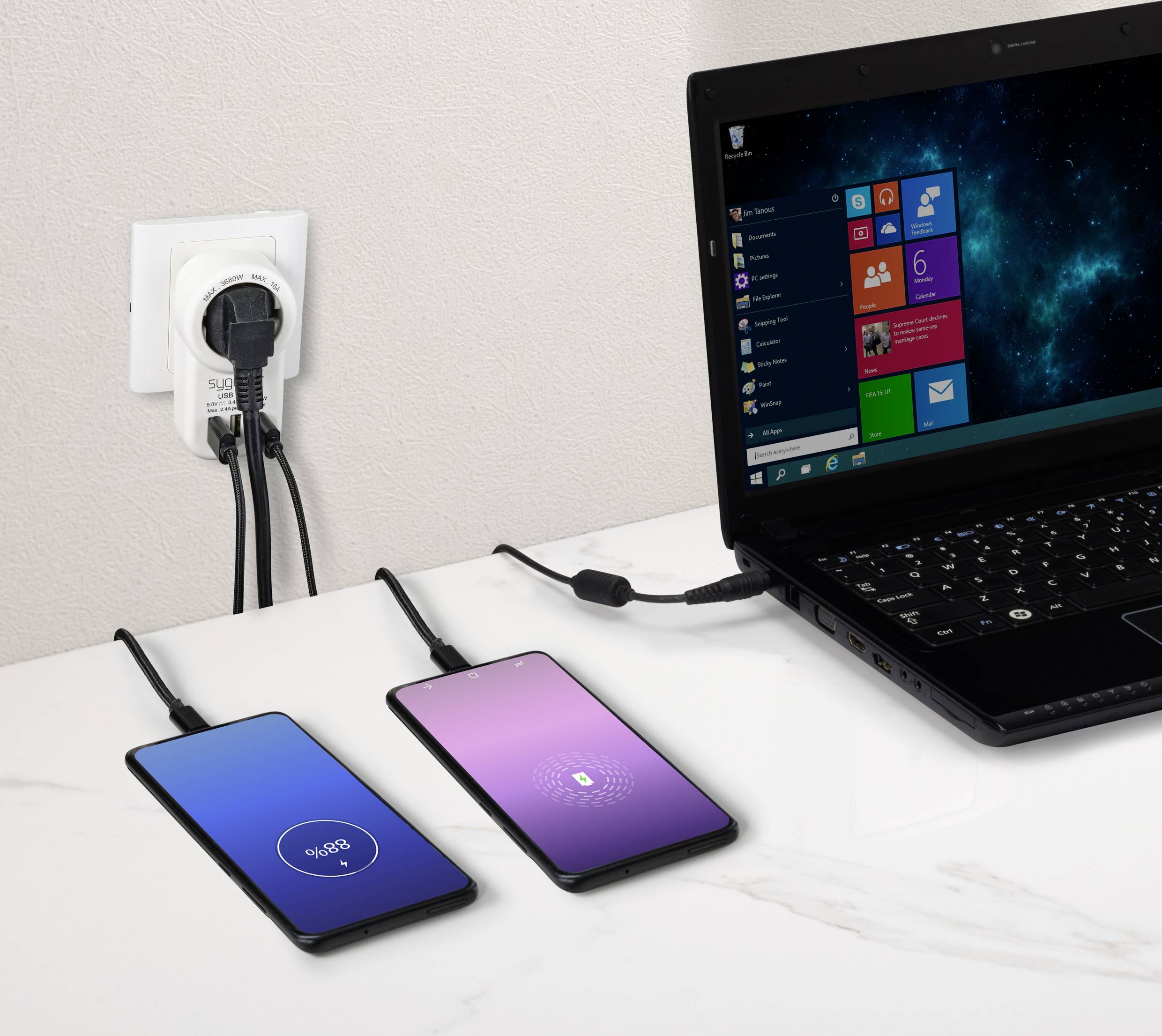 Egy fekete laptop Windows-képernyővel, két töltődő okostelefon; az egyik 90%-os, a másik 18%-os töltöttséget mutat, a háttérben egy elektromos csatlakozó.