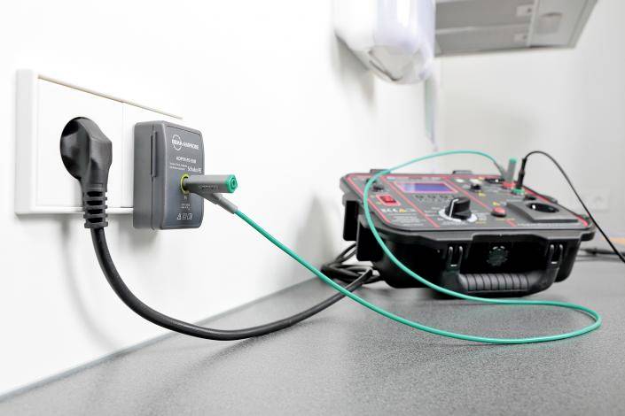 Egy tesztelőkészülék csatlakoztatva van egy fali aljzathoz. Az elektromos biztonságot és teljesítményt teszteli, amelyet kábelek és mérőműszerek mutatnak.