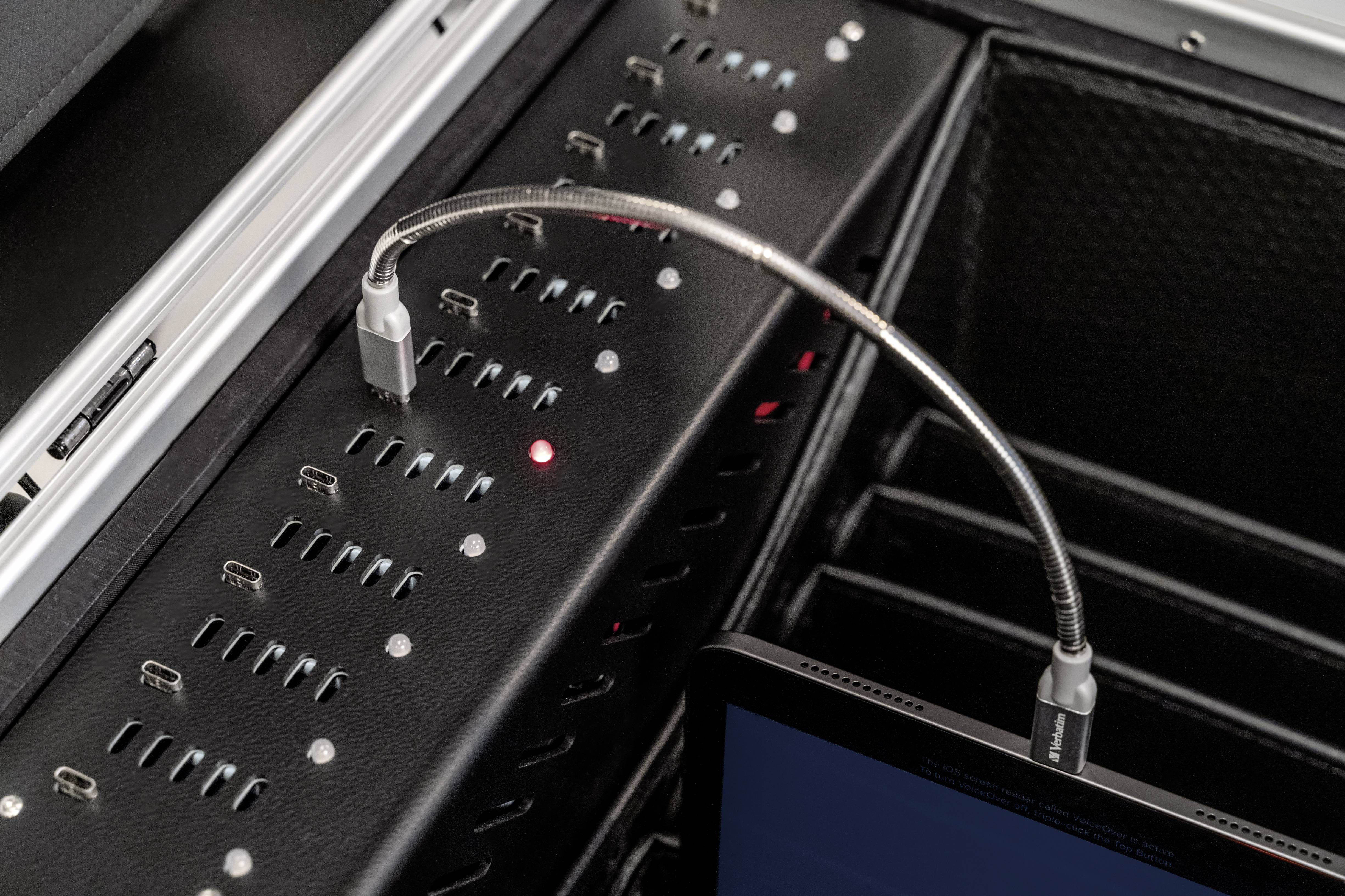 Több csatlakozóval rendelkező töltőállomás, piros fény jelzi az aktív töltési folyamatot. Egy rugalmas kábel köti össze a készüléket a töltőállomással.