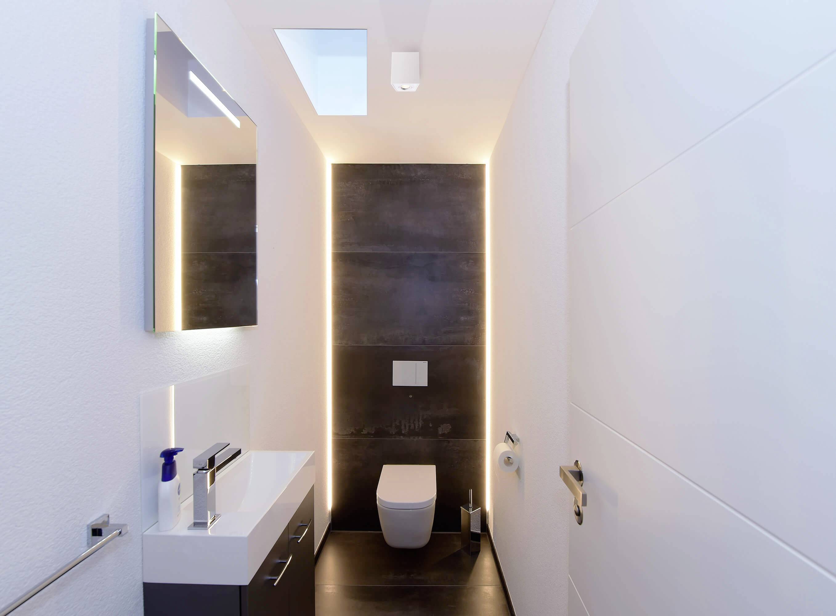 Modern fürdőszoba fali WC-vel, mosdókagylóval és tükörrel. Diszkrét világítás hangsúlyozza a fekete csempés hátteret.