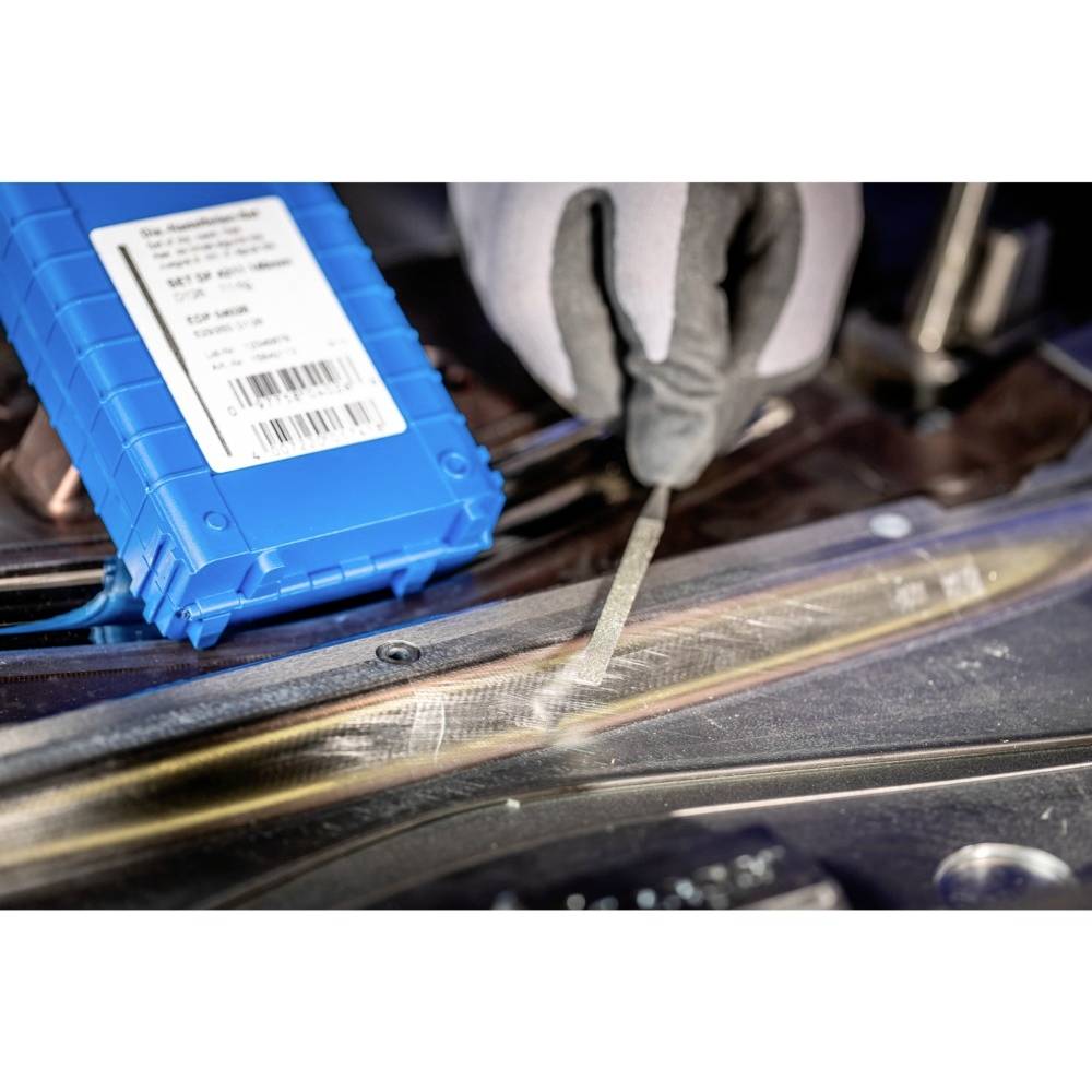 Egy személy kesztyűben reszel egy fémdarabot. A háttérben egy kék alkatrész fekszik egy címkével.