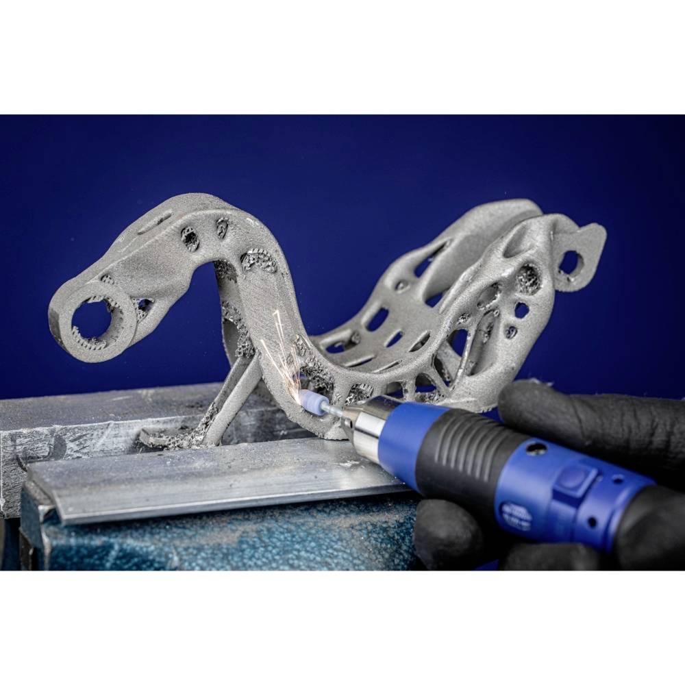 Egy kesztyűs kéz egy komplex, fémes 3D-nyomtatott tárgyat munkál meg egy kék szerszámmal.