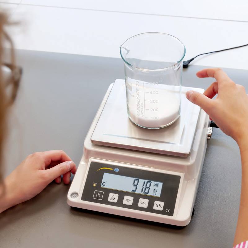 Egy kéz mérőhengert helyez folyadékkal egy digitális mérlegre, amely 918,9 gramm súlyt jelez.