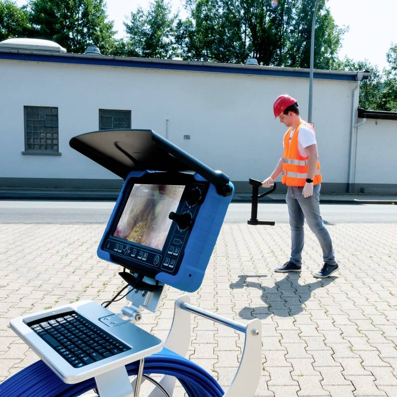Egy védősapkát és fényvisszaverő mellényt viselő technikus hordozható vizsgálókészüléket kezel egy épület előtti térburkolaton.