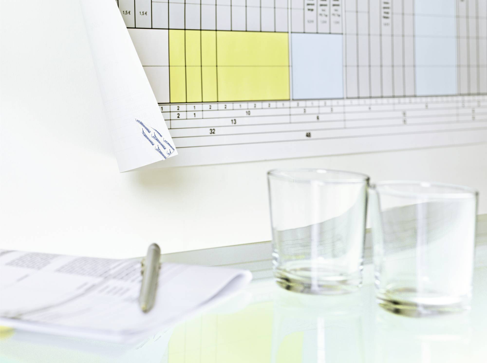 Két üres vizes pohár áll egy asztalon egy nyitott noteszblokk és toll mellett, a háttérben pedig egy fali naptár látható.