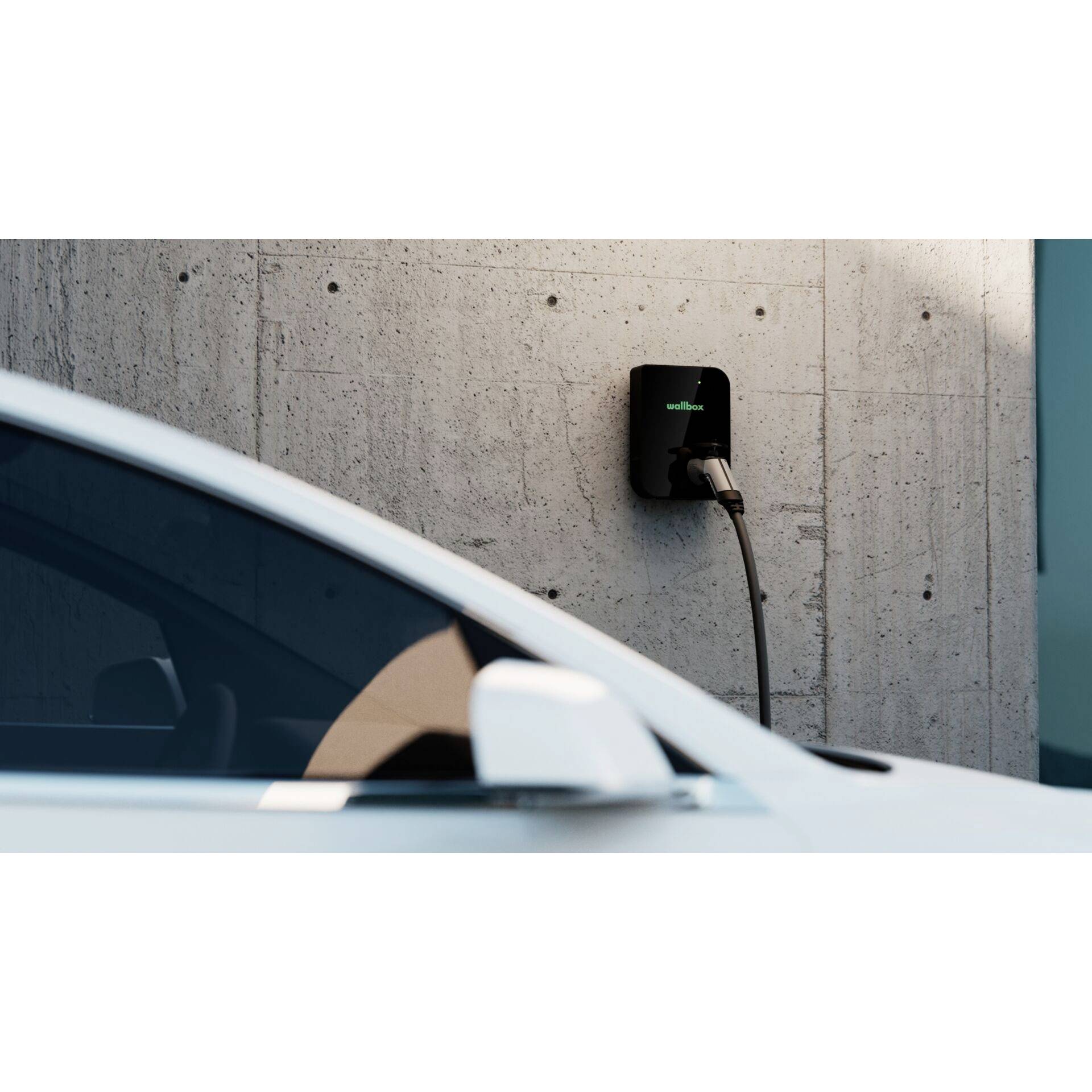 Egy elektromos autó egy betonfalon lévő töltőállomáson tölt. Az előtérben az autó oldalsó tükre látható.