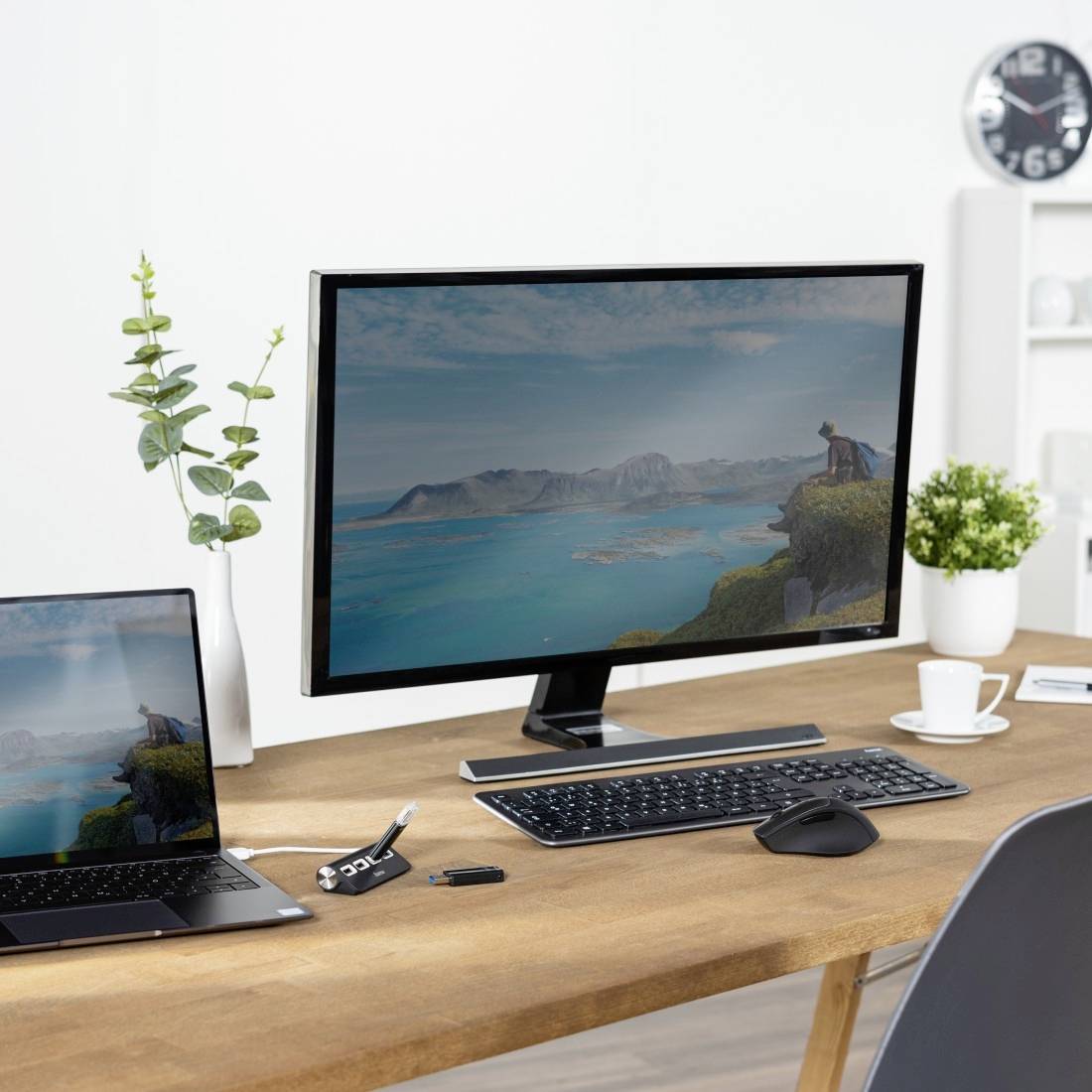 Két monitor áll az íróasztalon, amelyek egy tengeri tájat mutatnak hegyek között. Mellettük egy billentyűzet, egér és egy laptop helyezkedik el.
