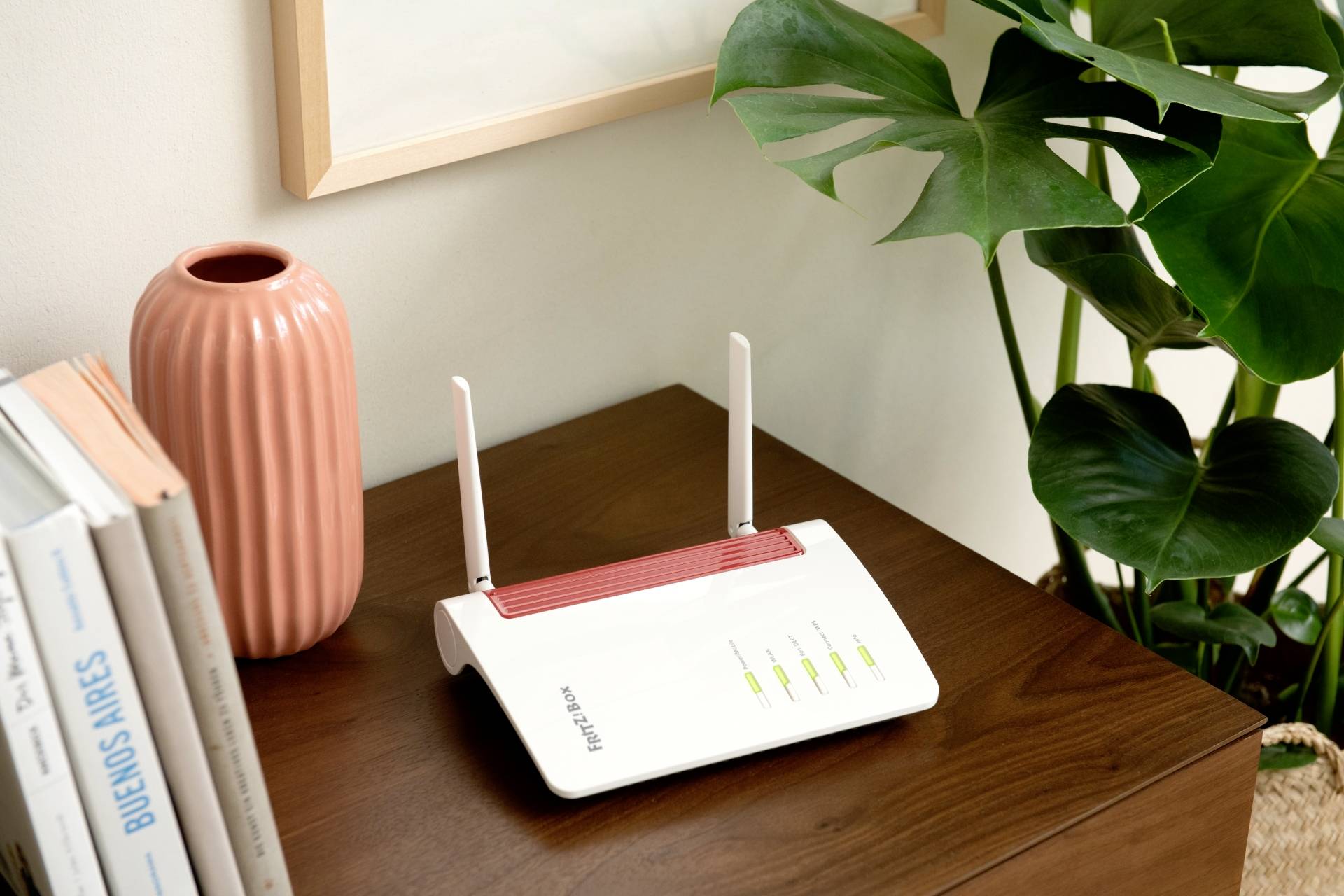 Egy fehér router négy világító LED-del és antennákkal egy faasztalon könyvek és egy növény mellett áll.