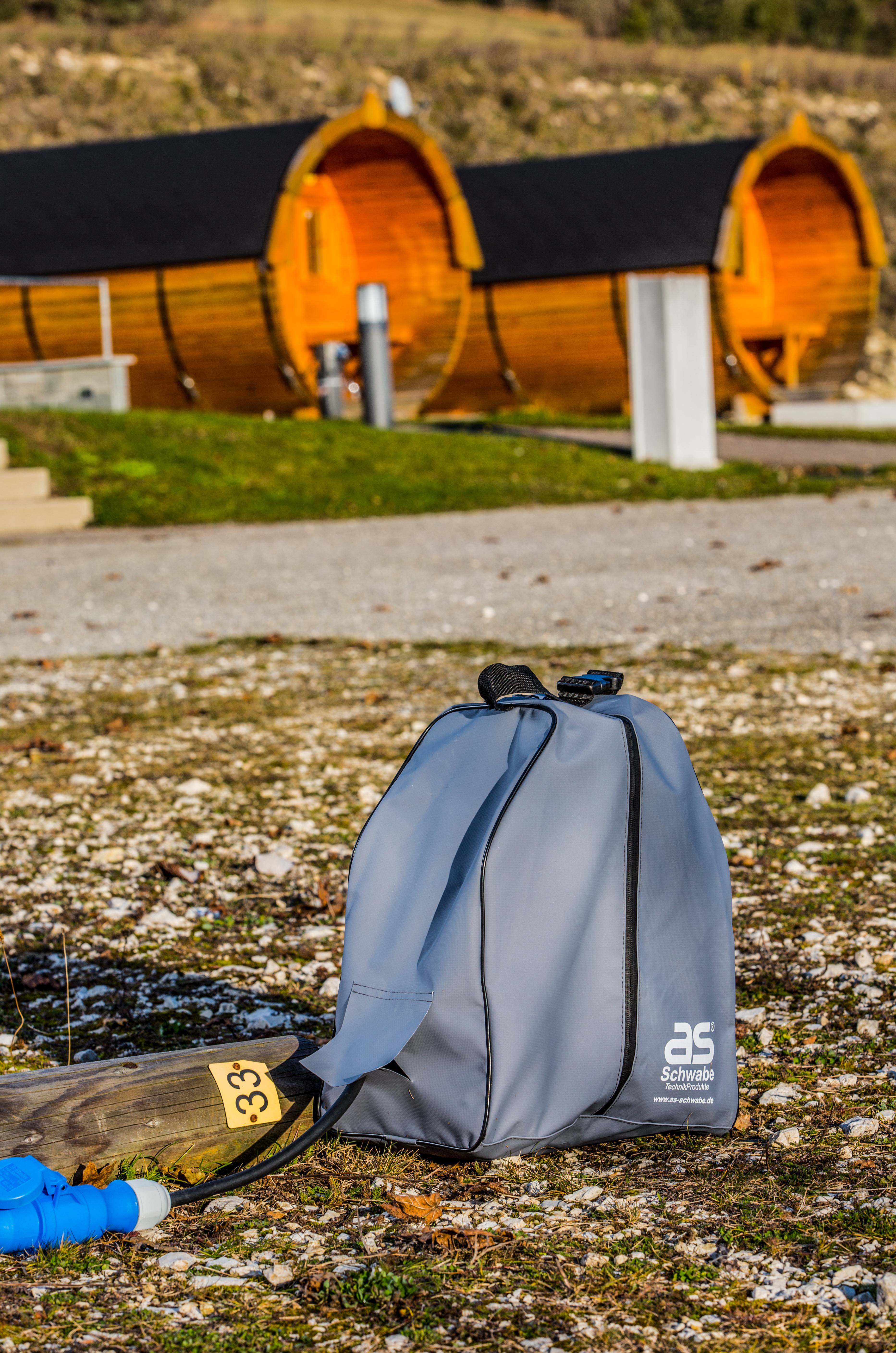 Egy szürke sporttáska áll egy kavicsos úton az előtérben. A háttérben faházak íves tetővel egy mezőn láthatók.